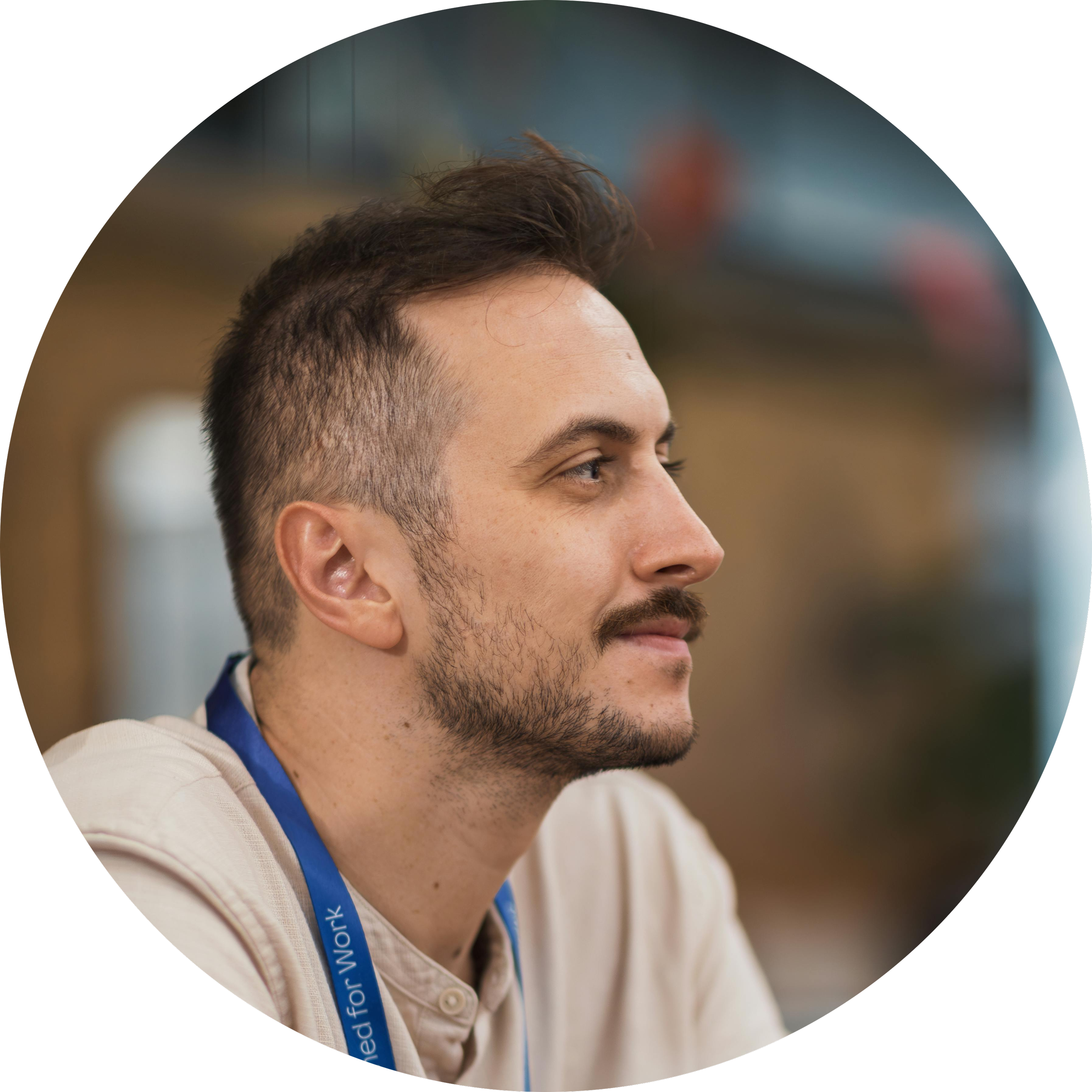 Side profile of a man with short, styled brown hair, a beard, and wearing a beige shirt with a blue lanyard around his neck.