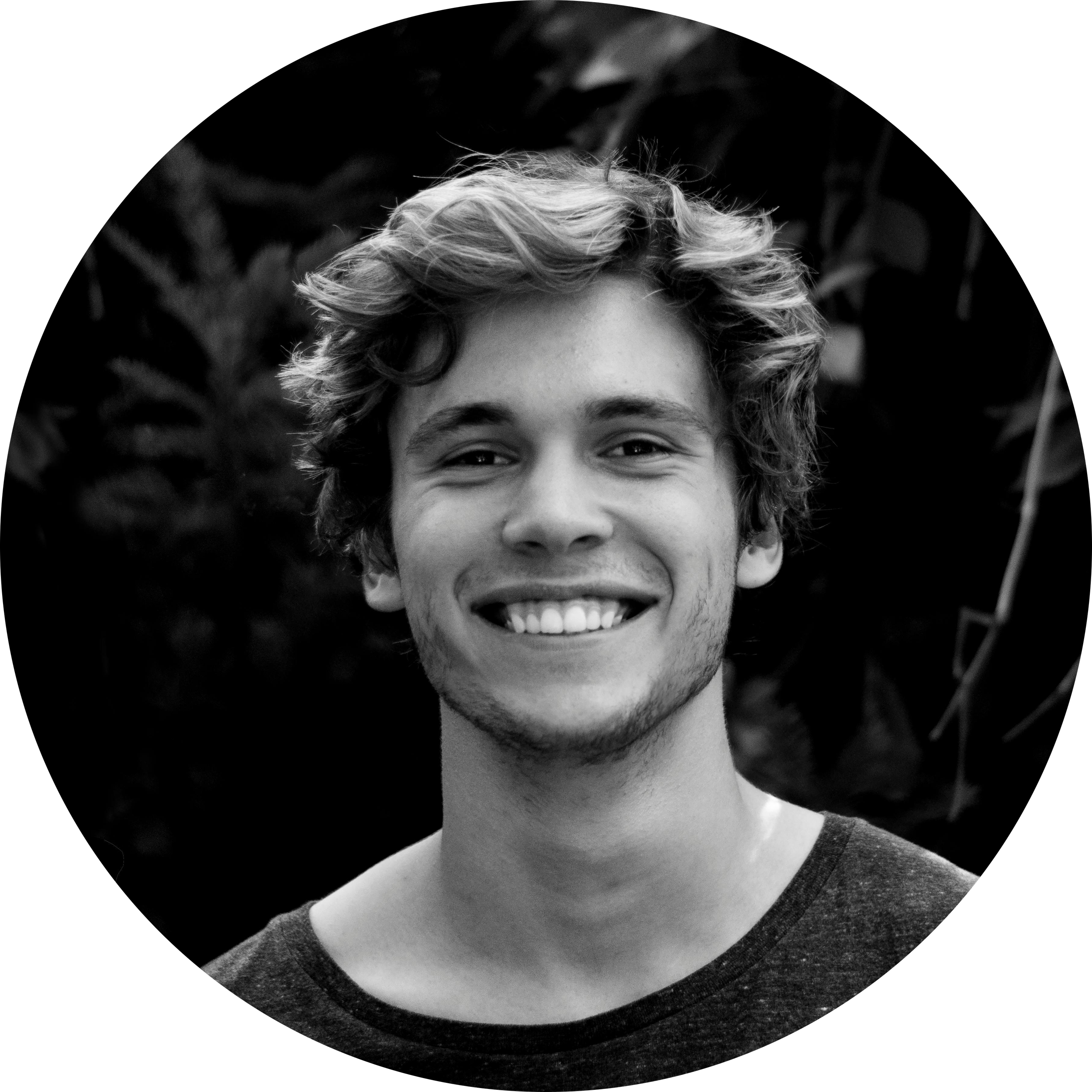 Black and white photo of a smiling young man with curly hair, wearing a casual shirt, outdoors with blurred foliage in the background.