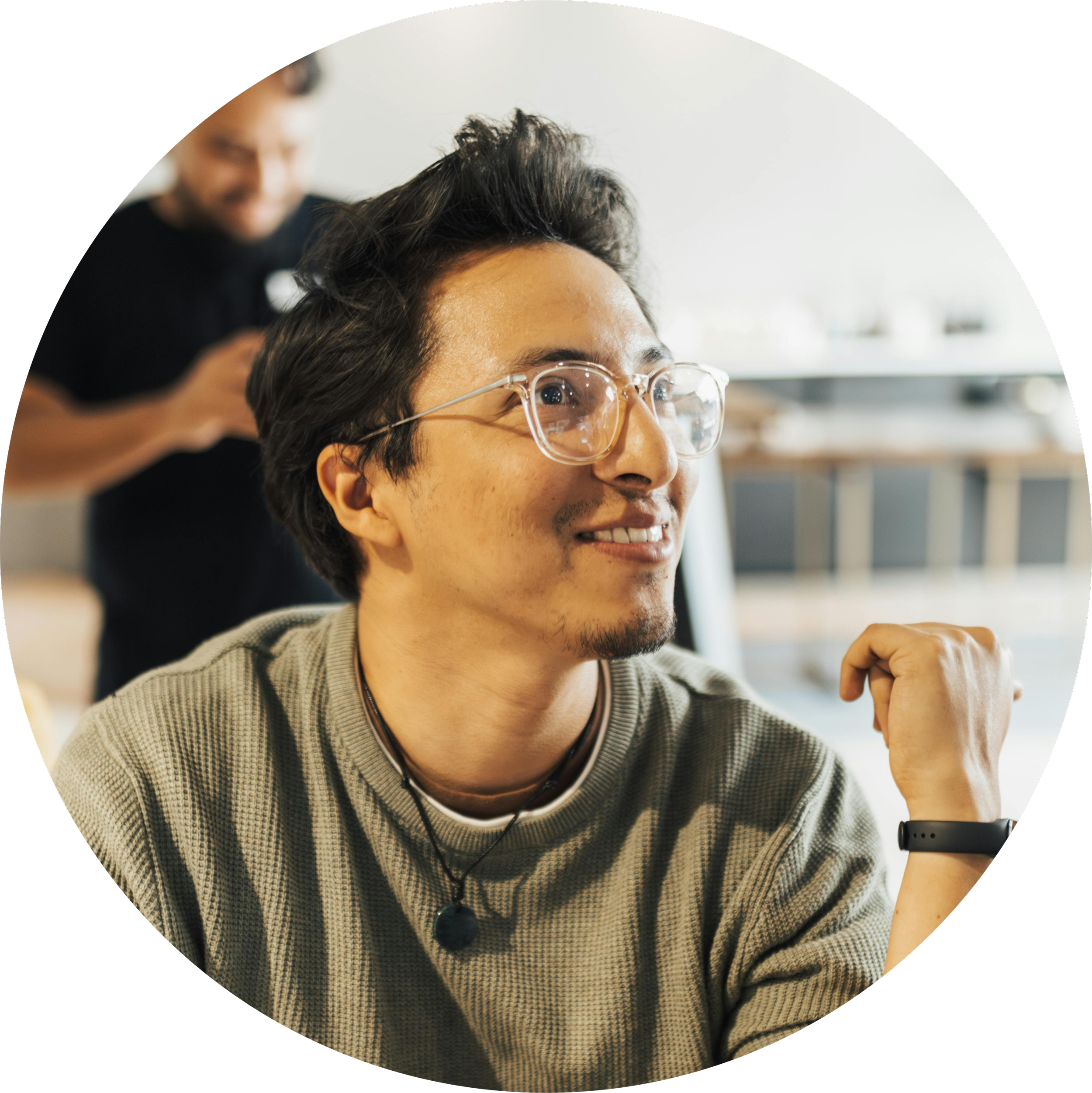 A young man wearing glasses and a beige sweater smiling, with a man in the background looking at a phone.