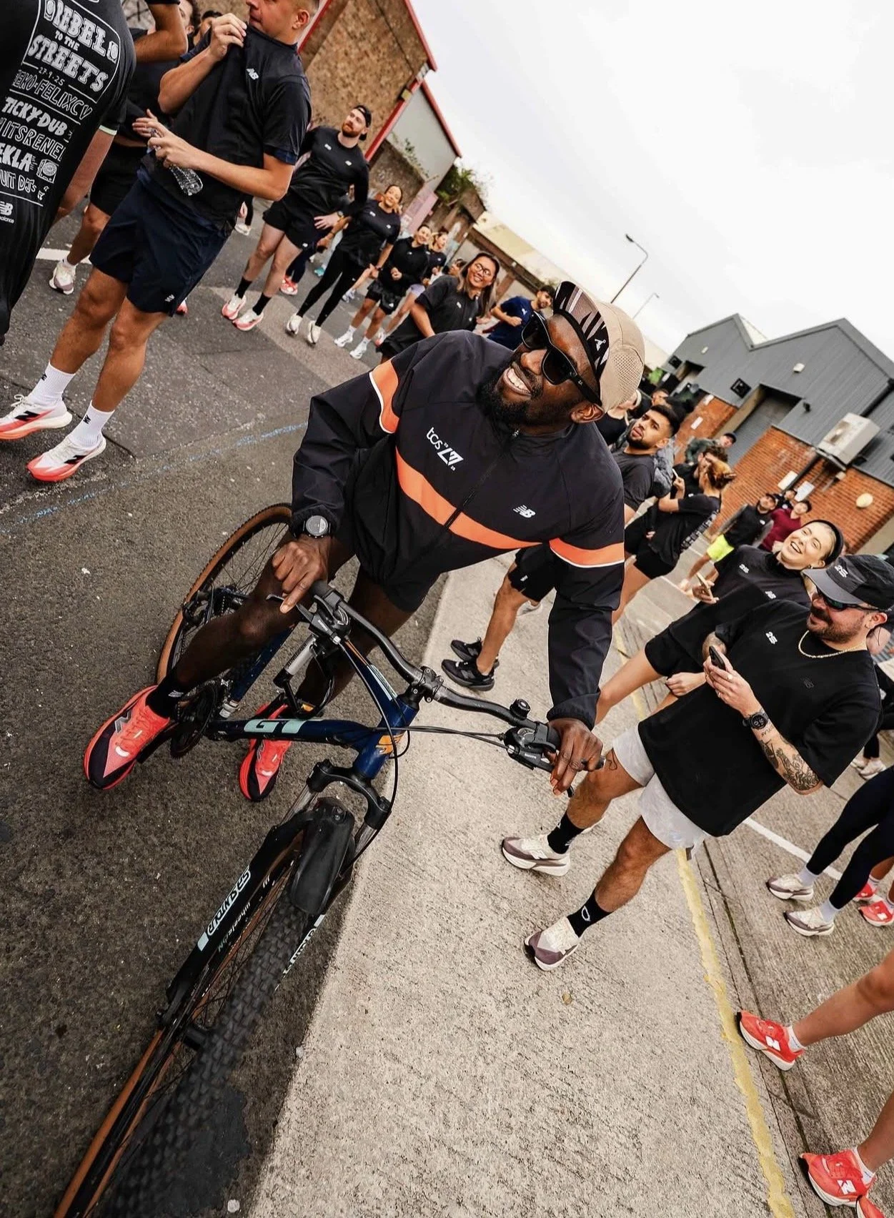 Man smiling and riding a mountain bike outdoors, surrounded by a group of people in athletic clothing, walking and chatting on a cloudy day.