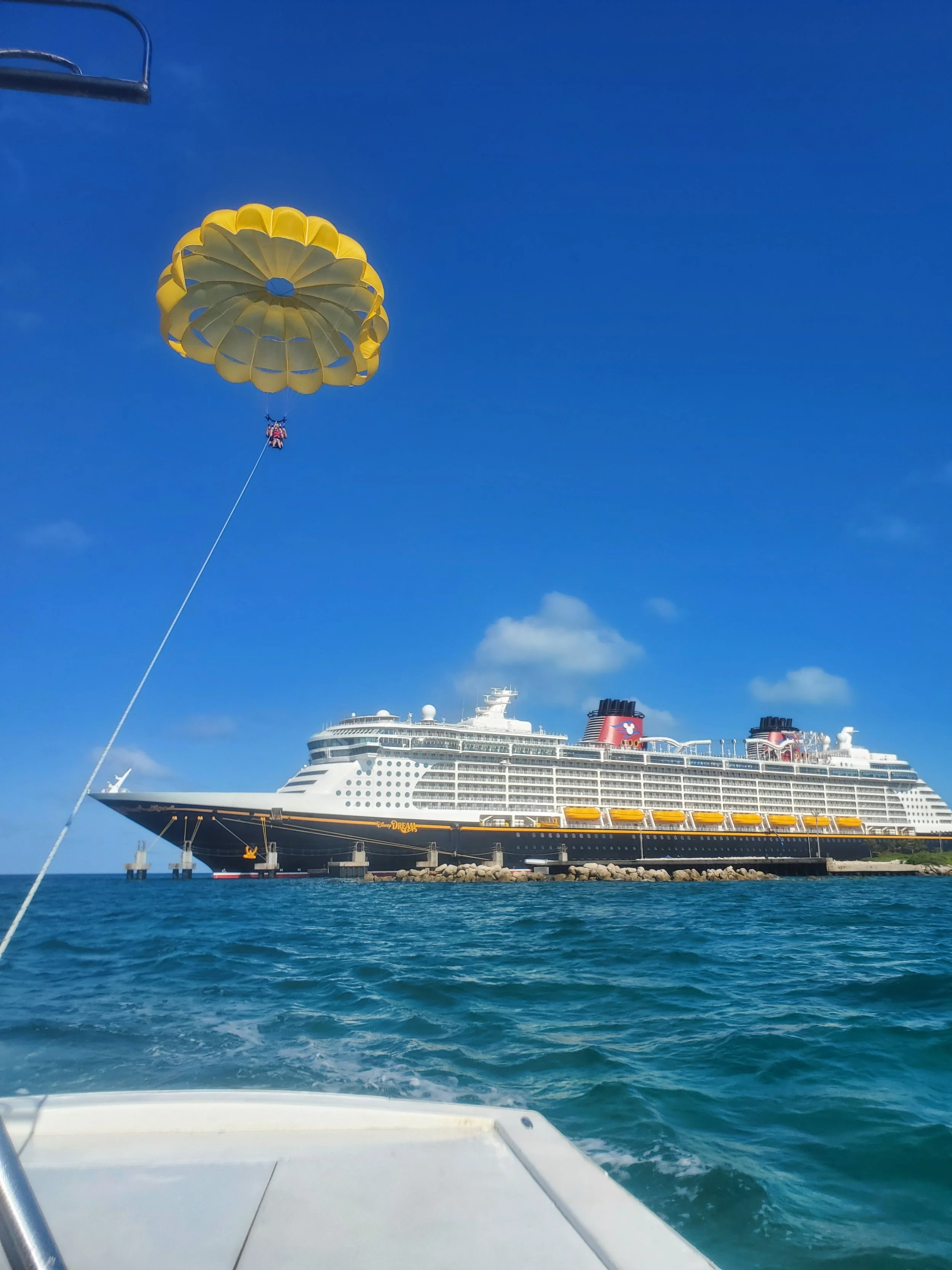 A large cruise ship docked at a port with bright blue sky, visible whale tail, and a parasail with a person attached floating above.