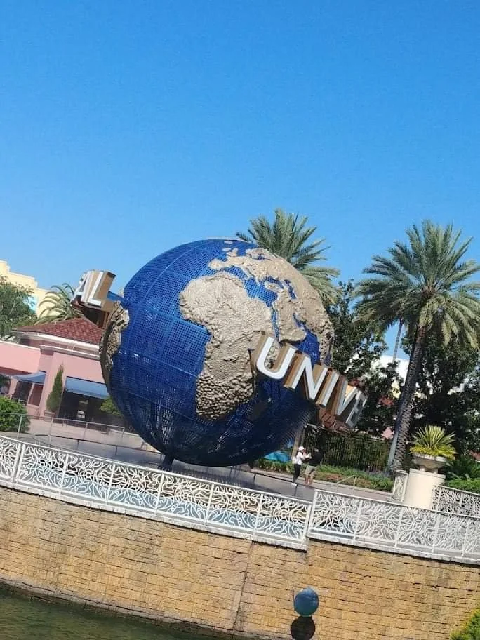 Large globe sculpture with 'UNIVERSAL' text, situated outdoors near water, with palm trees and buildings in the background.
