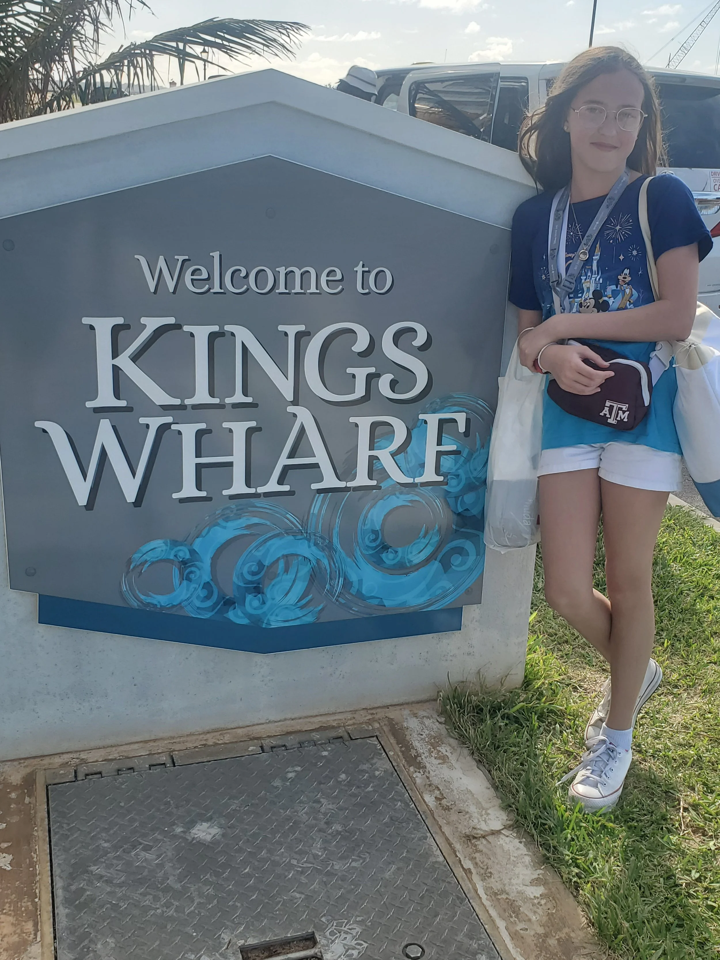 A young woman leaning on a sign that says ‘Welcome to Kings Wharf’ with a background of parked cars and palm trees.
