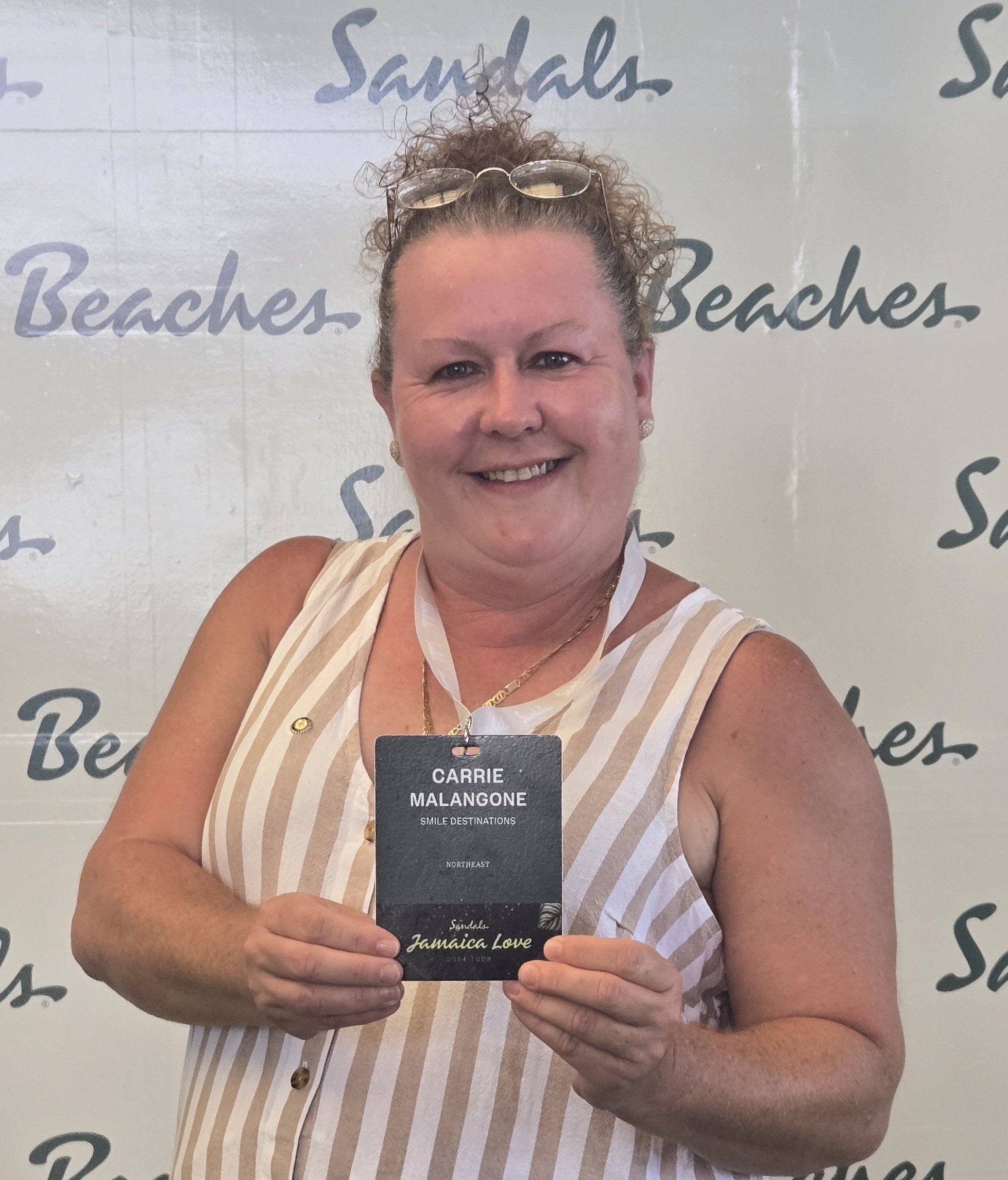 A woman with curly hair, wearing glasses on her head, a striped sleeveless top, and jewelry, holding a name badge that reads 'Carrie Malangone, Smile Destinations, Northeast, Jamaica Love, 2024 Tour', standing in front of a backdrop with the word 'Sandals Beaches' repeatedly printed.
