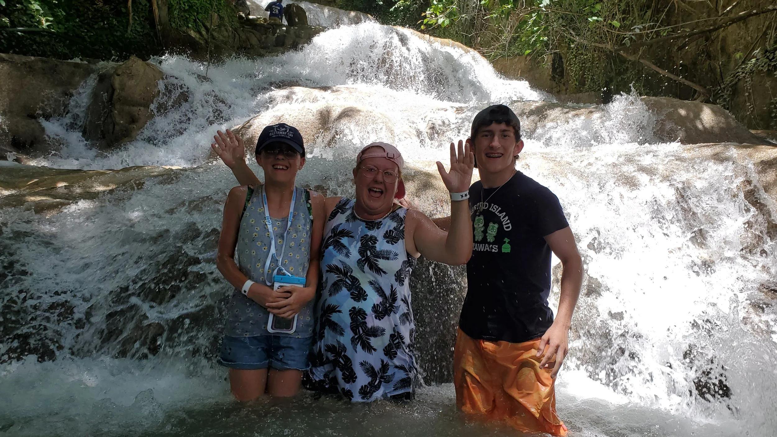 Three smiling people standing in a river with a waterfall in the background, all waving at the camera. The group includes a woman with glasses and a pink cap, a young woman with a blue tank top and a hat, and a young man in a black T-shirt and orange shorts.