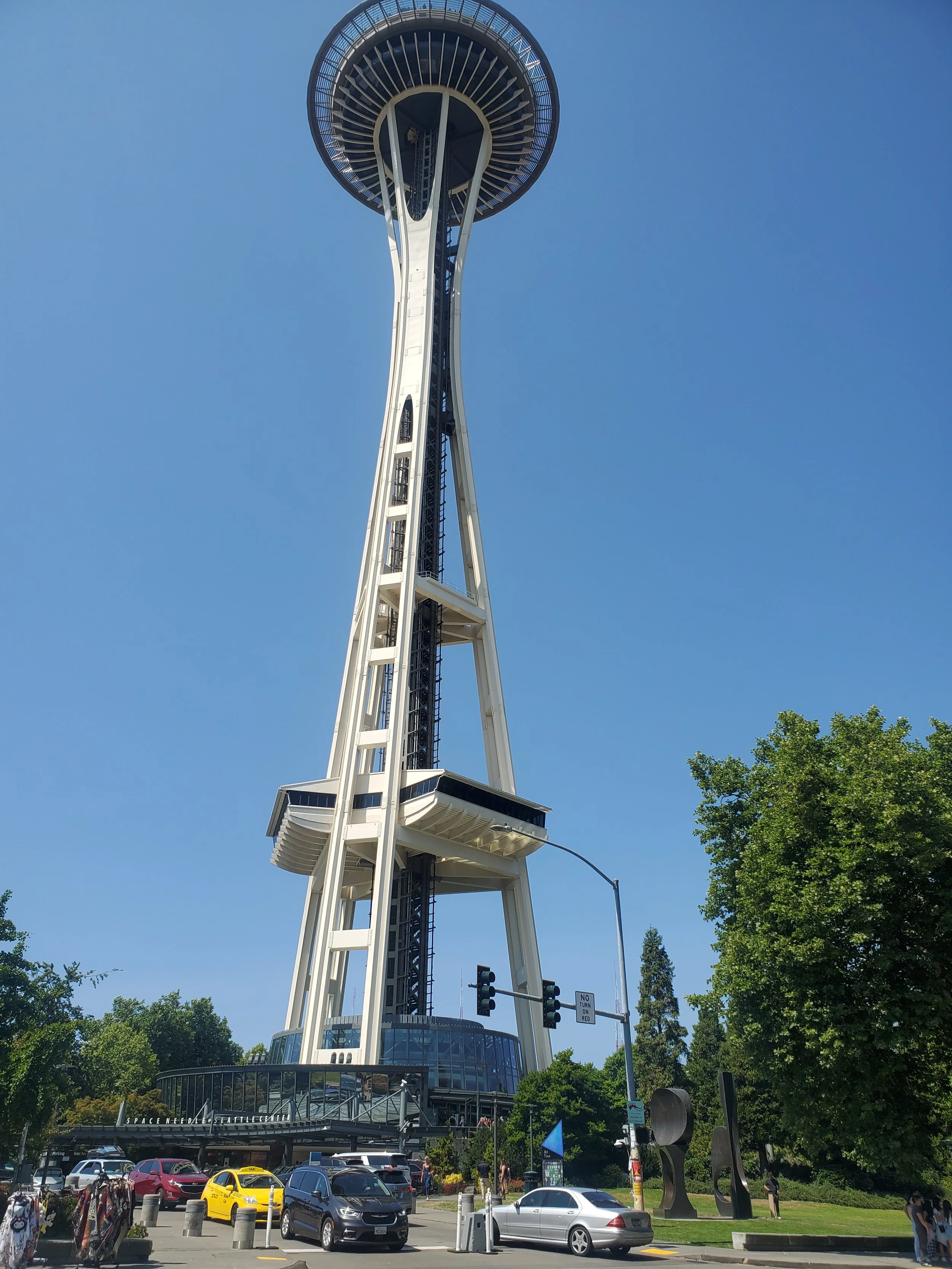 Seattle Space Needle with blue sky, trees, cars, and people in foreground.