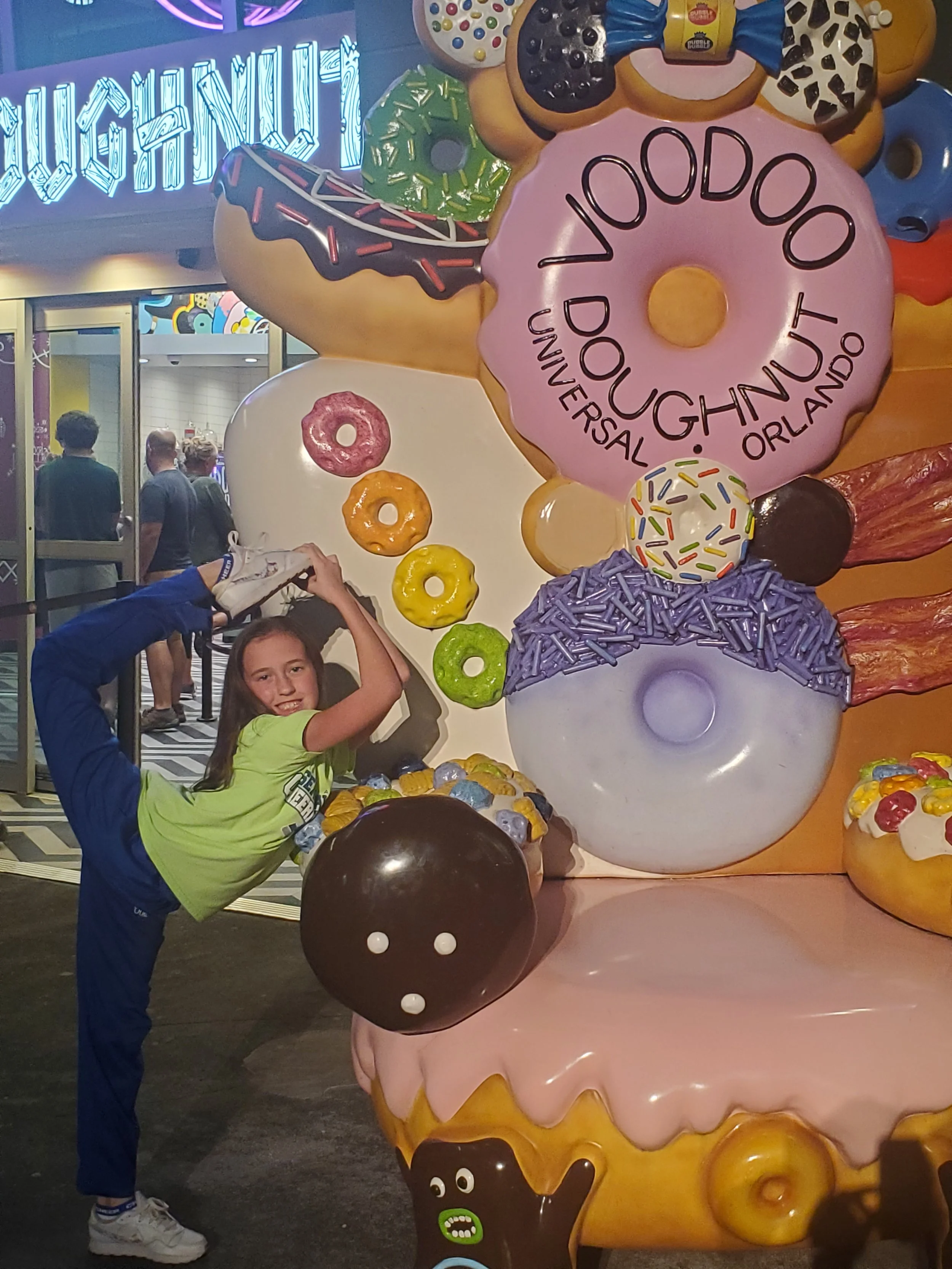A young girl in a bright green shirt and blue pants poses with a playful kick and holding her foot above her head, standing next to a large colorful display of oversized doughnuts with various icing and sprinkles at an amusement park.