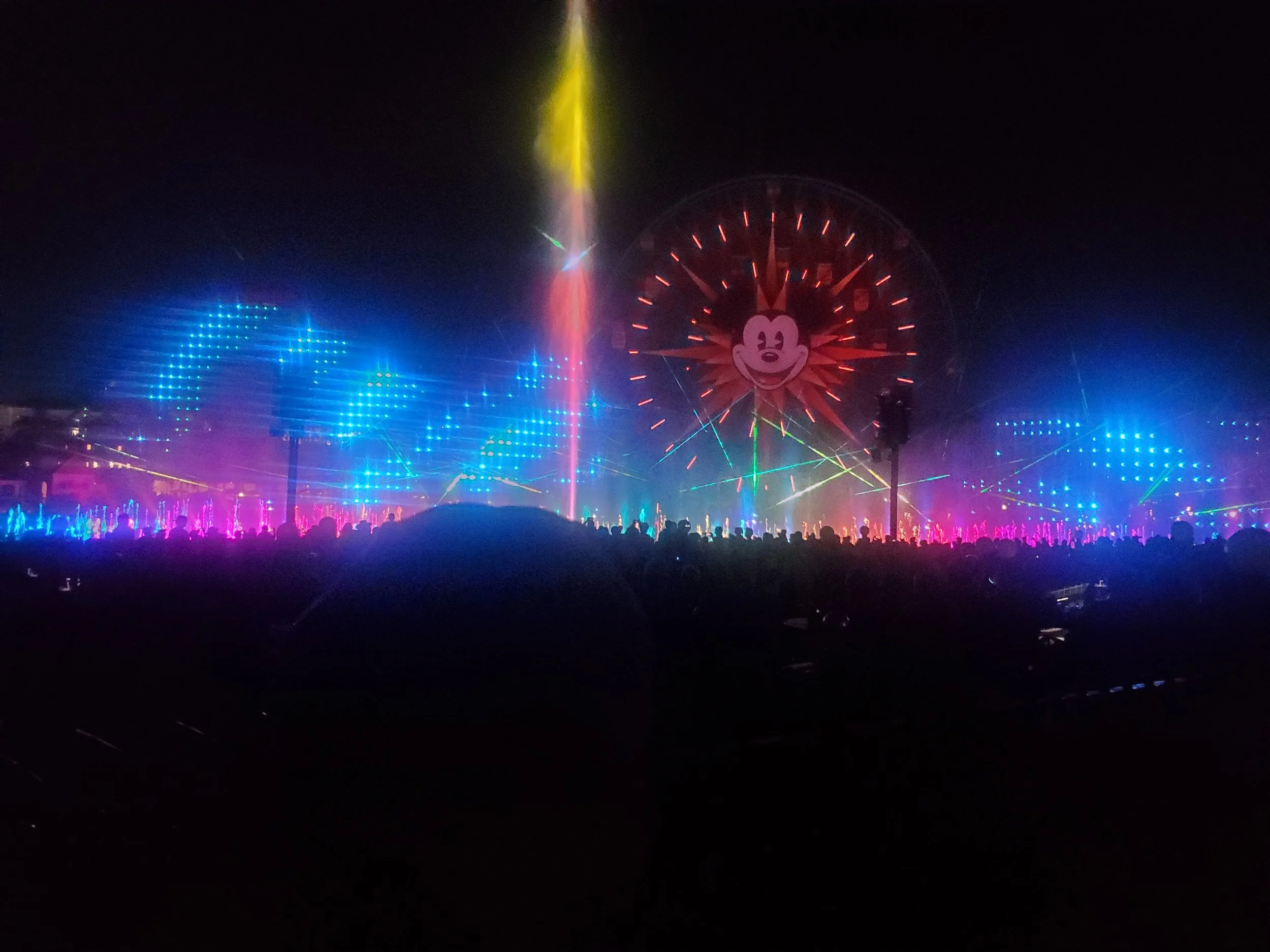 Nighttime scene at a Disney theme park with colorful lights, lasers, and a large Ferris wheel featuring Mickey Mouse's face.