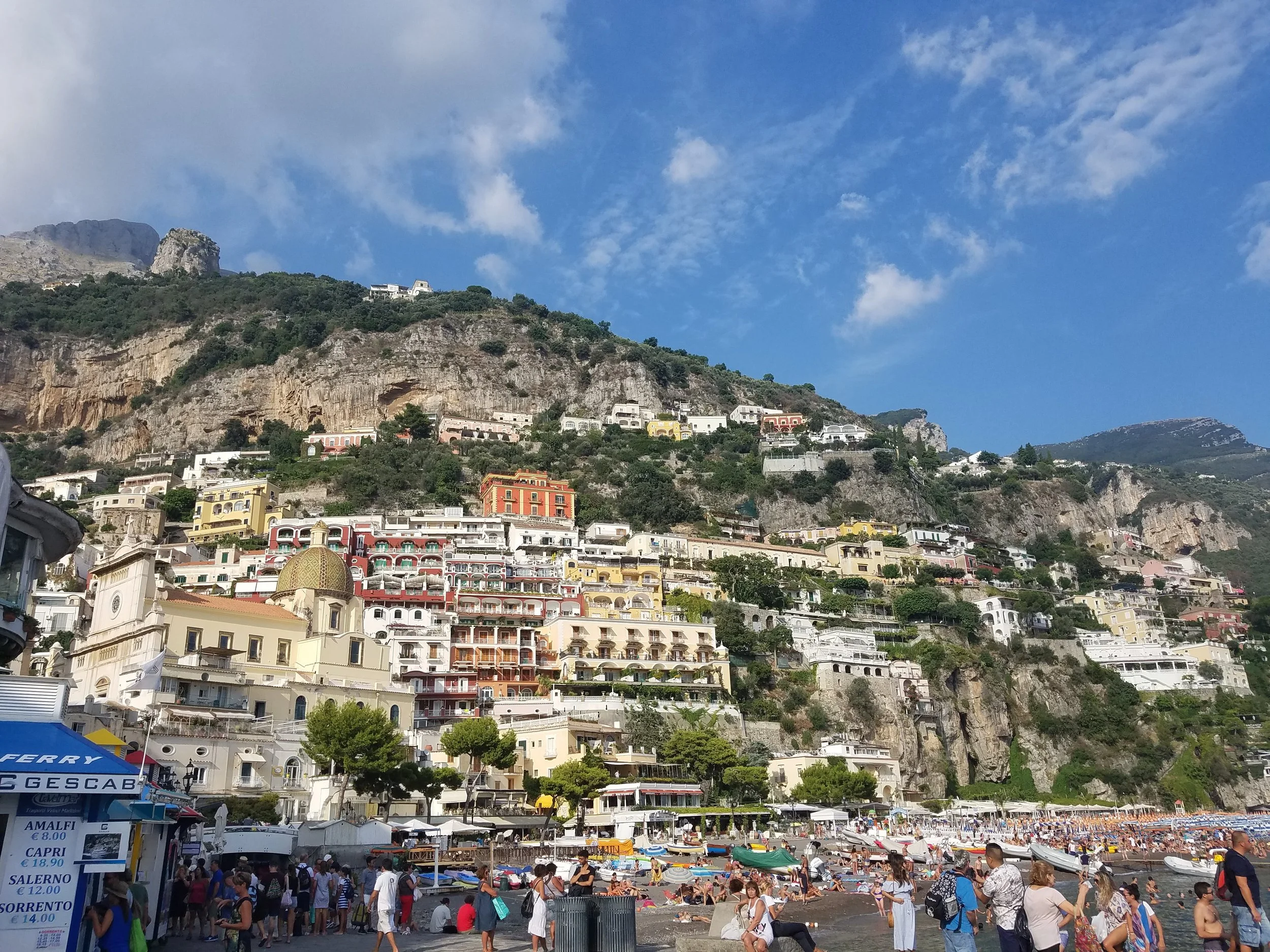 Colorful buildings on a hillside overlooking the beach in Positano, Italy, with people on the beach and boats in the water.