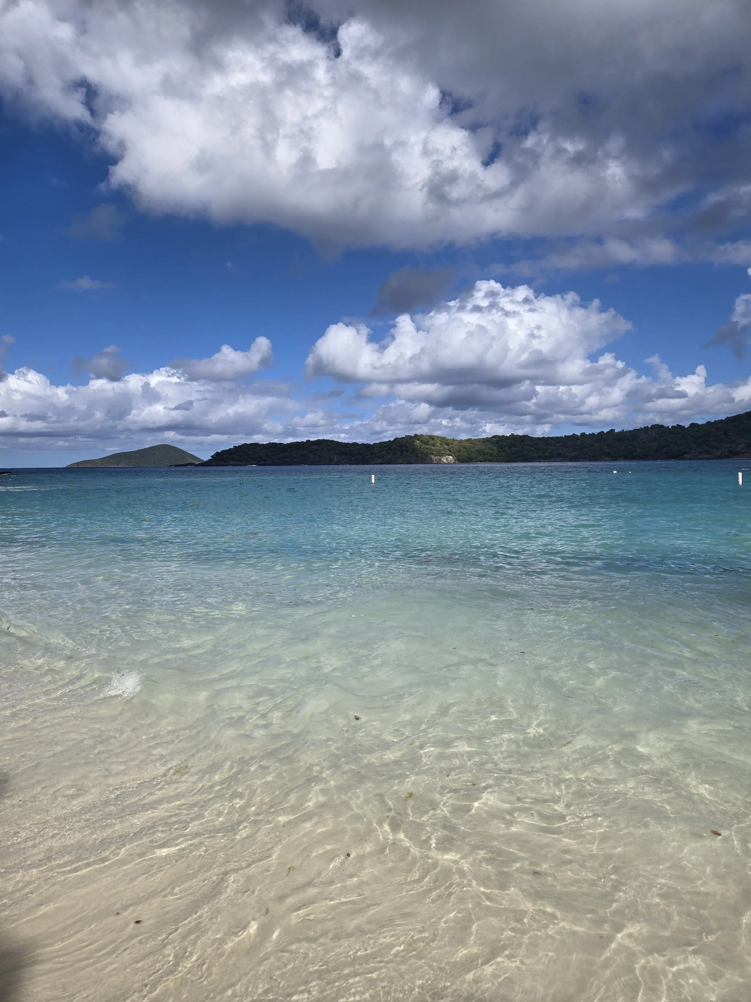 A beach scene with clear turquoise water, white sandy shore, and a background of green hills under a partly cloudy blue sky.
