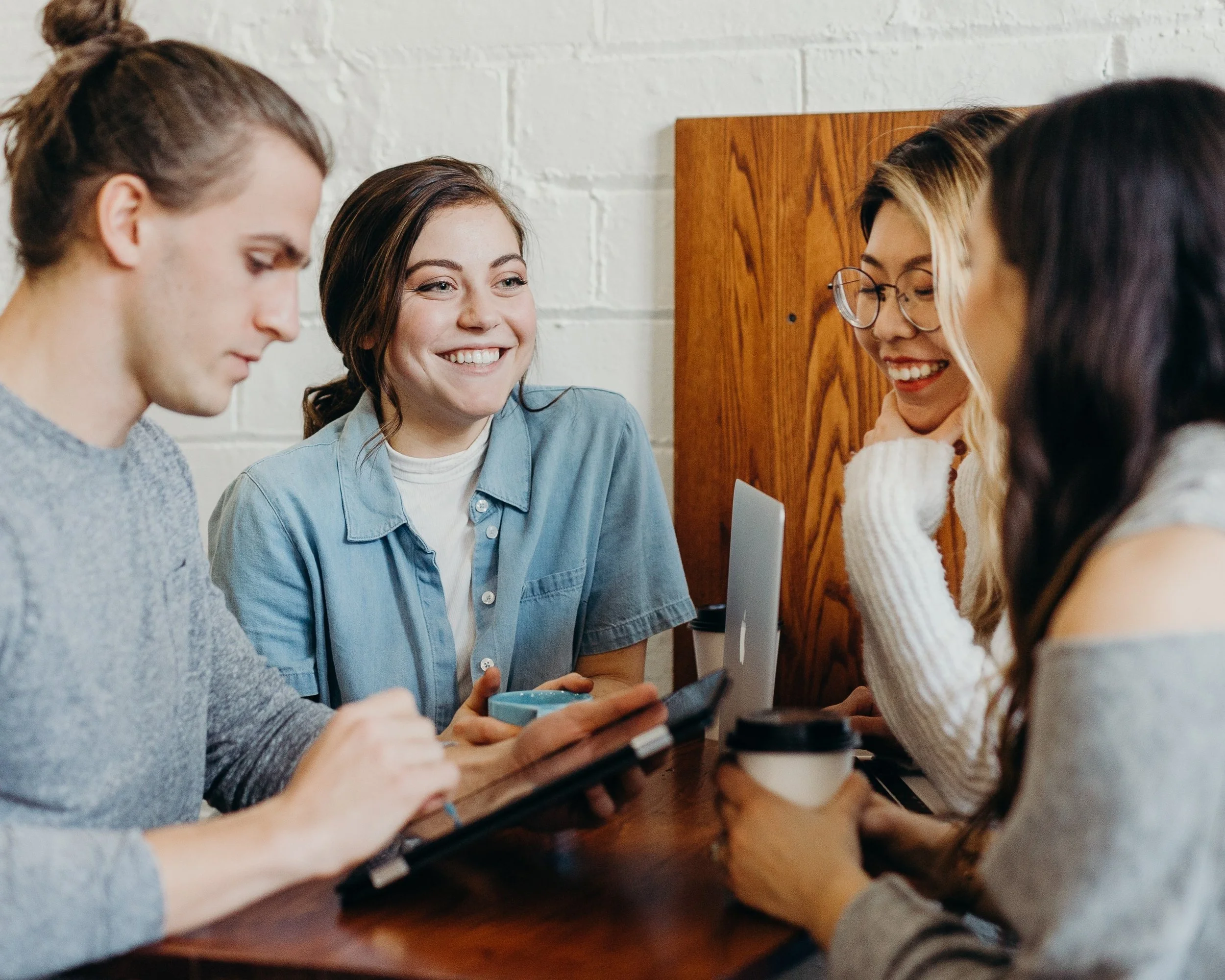 Four friends sitting at a table in a coffee shop, smiling and talking, with drinks and electronic devices on the table.