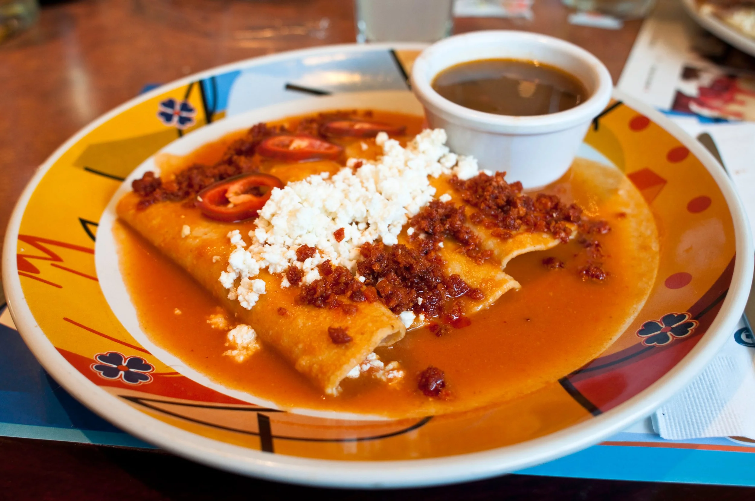 Plate of Mexican food with a cheese enchilada topped with tomato slices, crumbled cheese, and ground meat, served with a side of sauce in a small white cup.