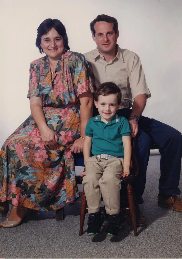 A family of three posing for a portrait; a woman in a colorful floral dress, a man in a beige shirt, and a young boy in a teal polo and khakis.