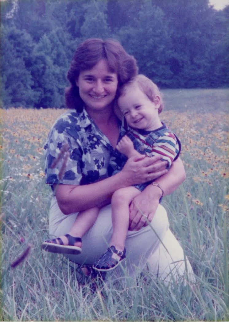 A woman with short brown hair smiling and holding a young boy with blonde hair in a field of tall grass and flowers, with trees in the background.