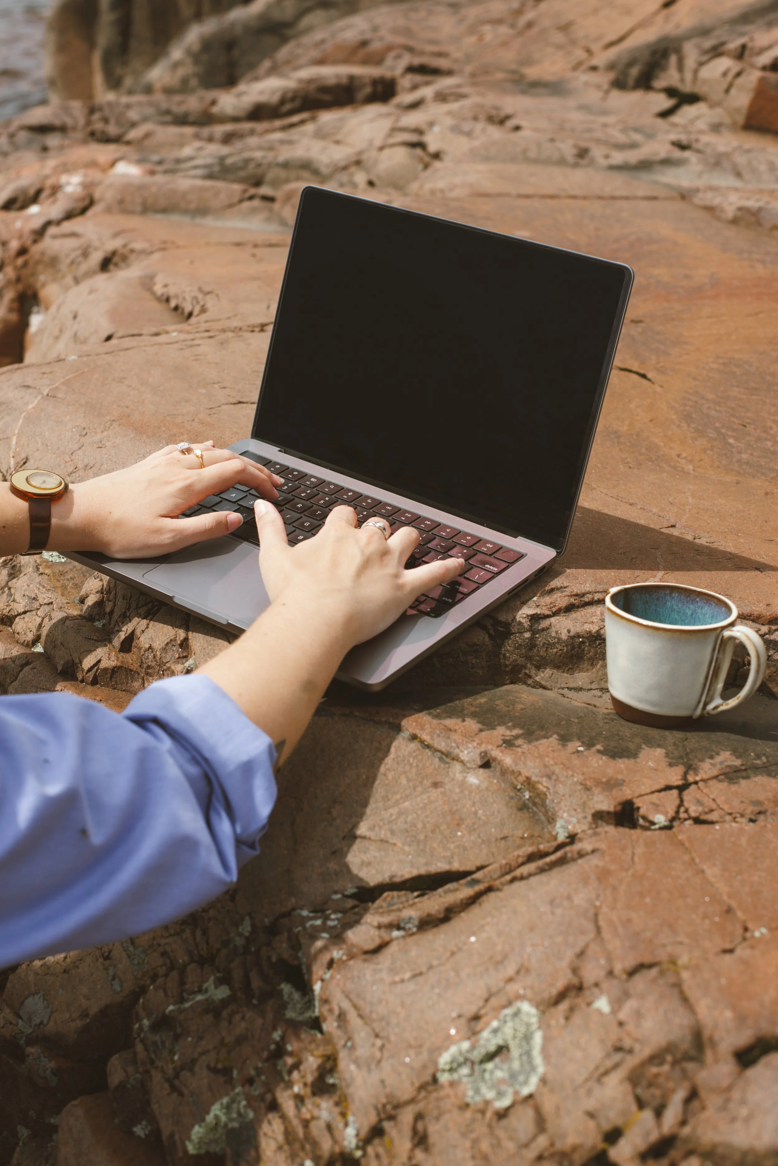 MacBook on seaside rocks with hands typing on keyboard and ceramic mug on the right side