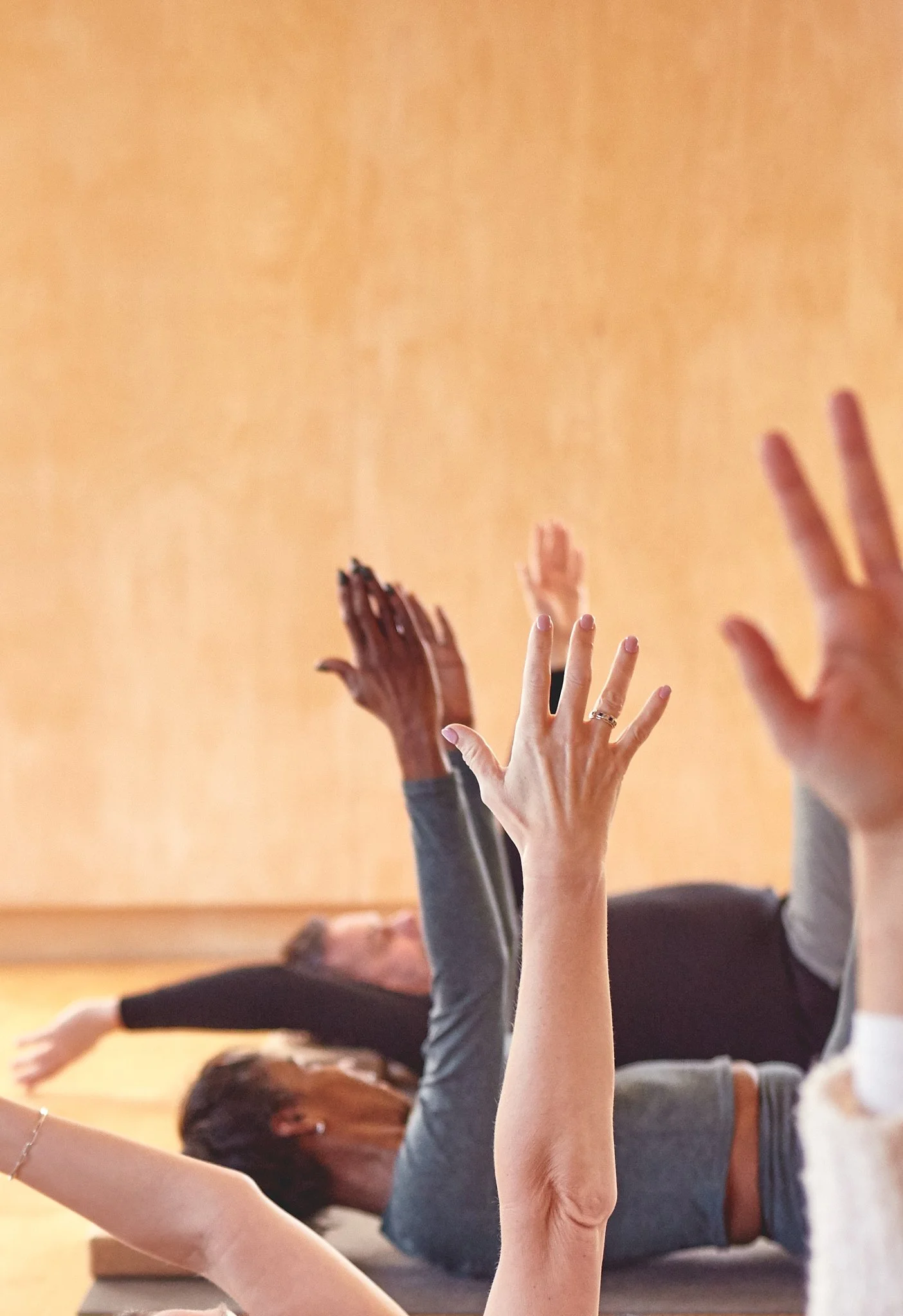People practicing pilates on mats, raising their hands during a class in a room with wooden floors and walls.
