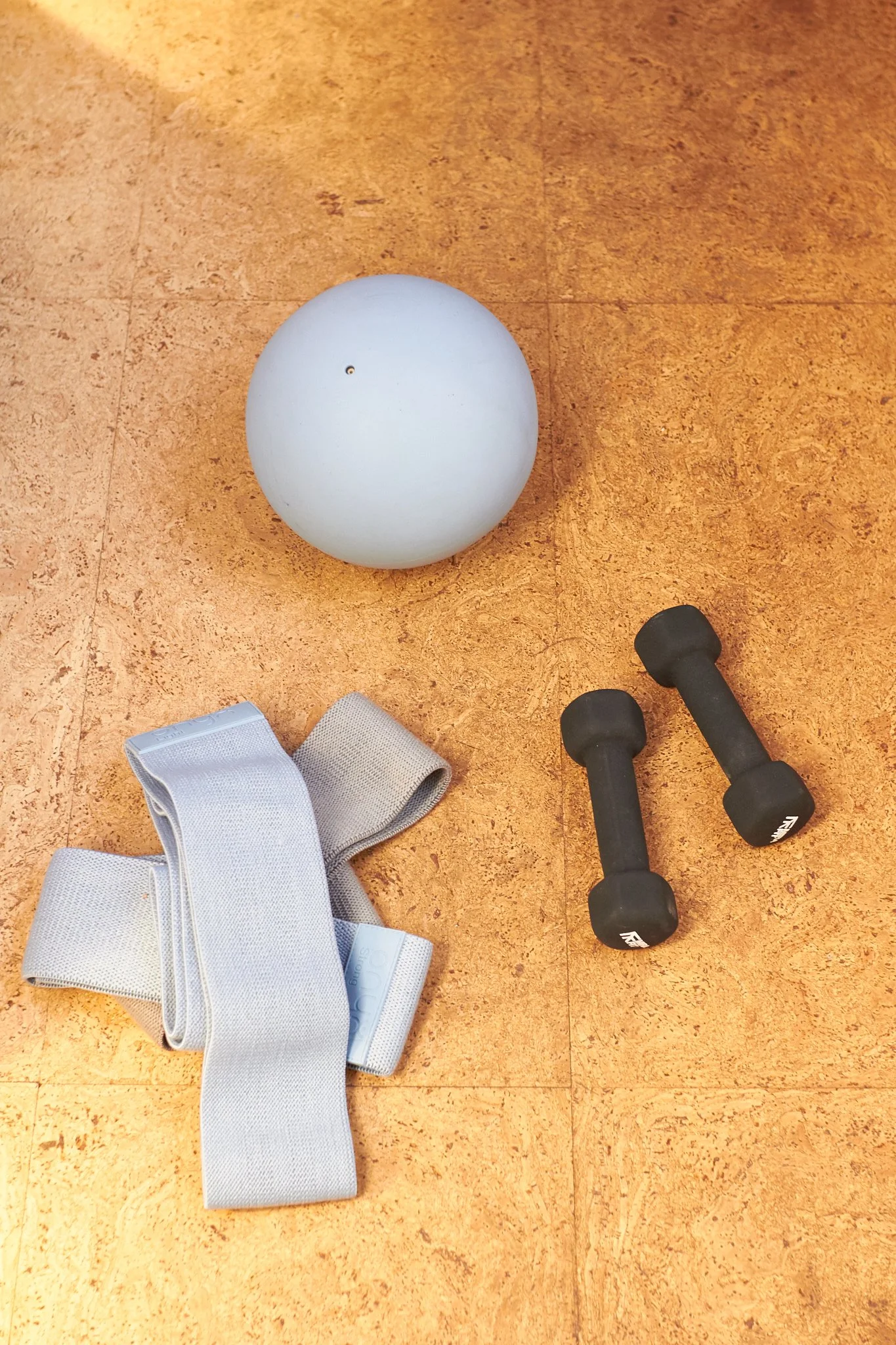 Fitness equipment including a large white exercise ball, a pair of small black dumbbells, and a gray resistance band on a brown floor.