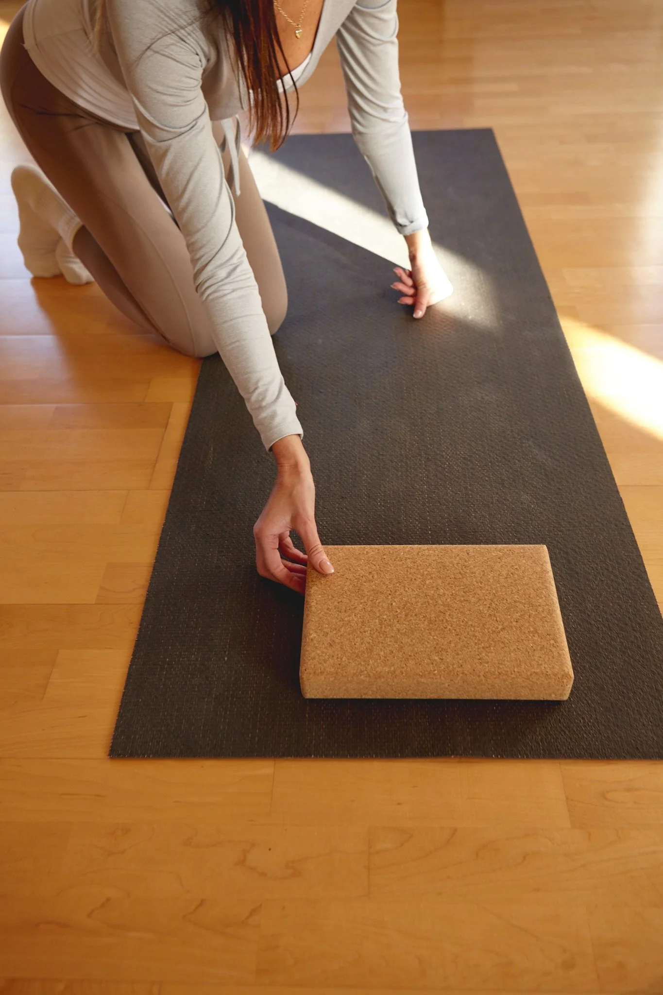 A person on a yoga mat, adjusting a cork yoga block while kneeling on the floor in a room with wooden flooring.