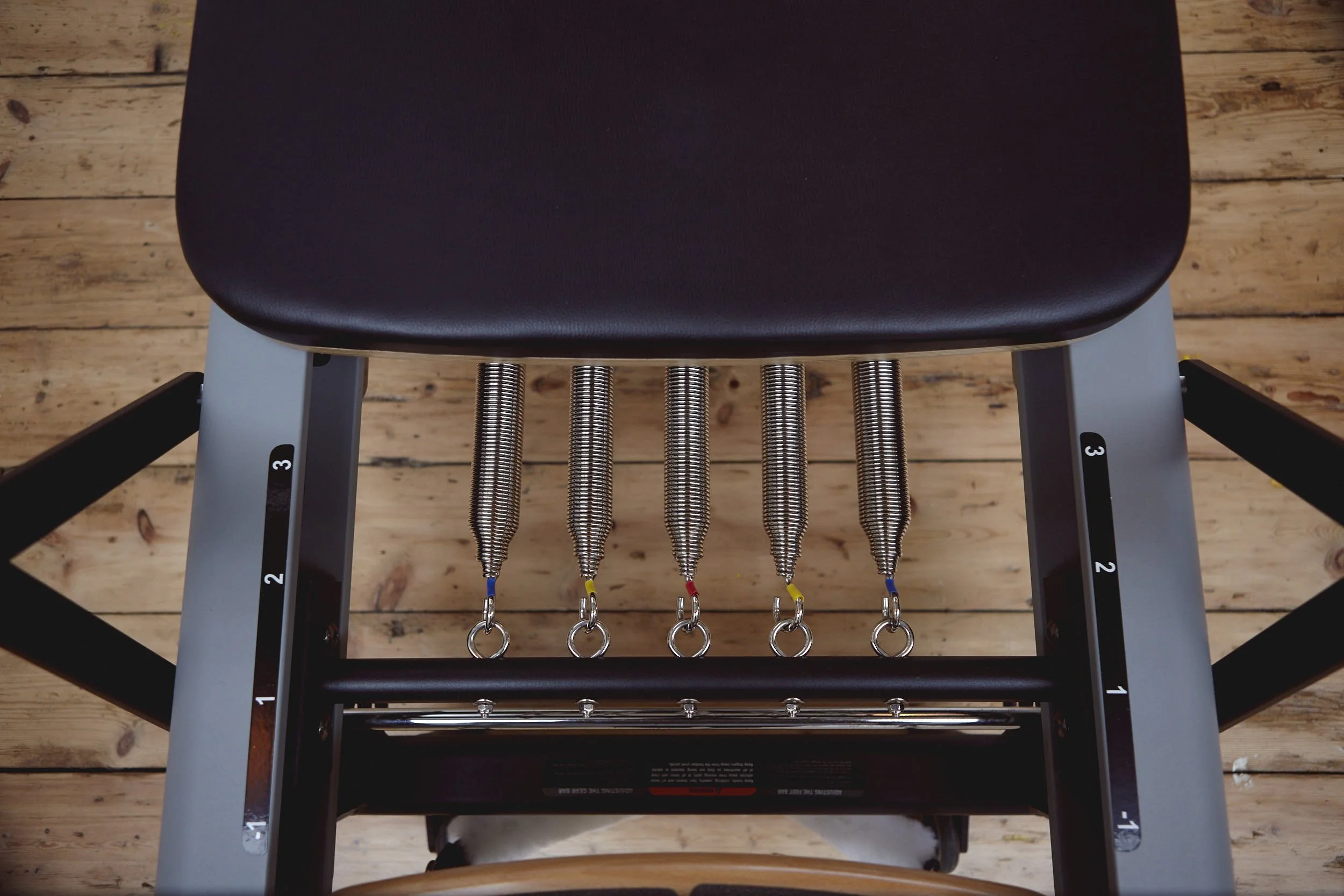 Six pendulum weights attached to springs hanging from a black bar on a Reformer Pilates machine, with a black padded seat and a wooden floor in the background.