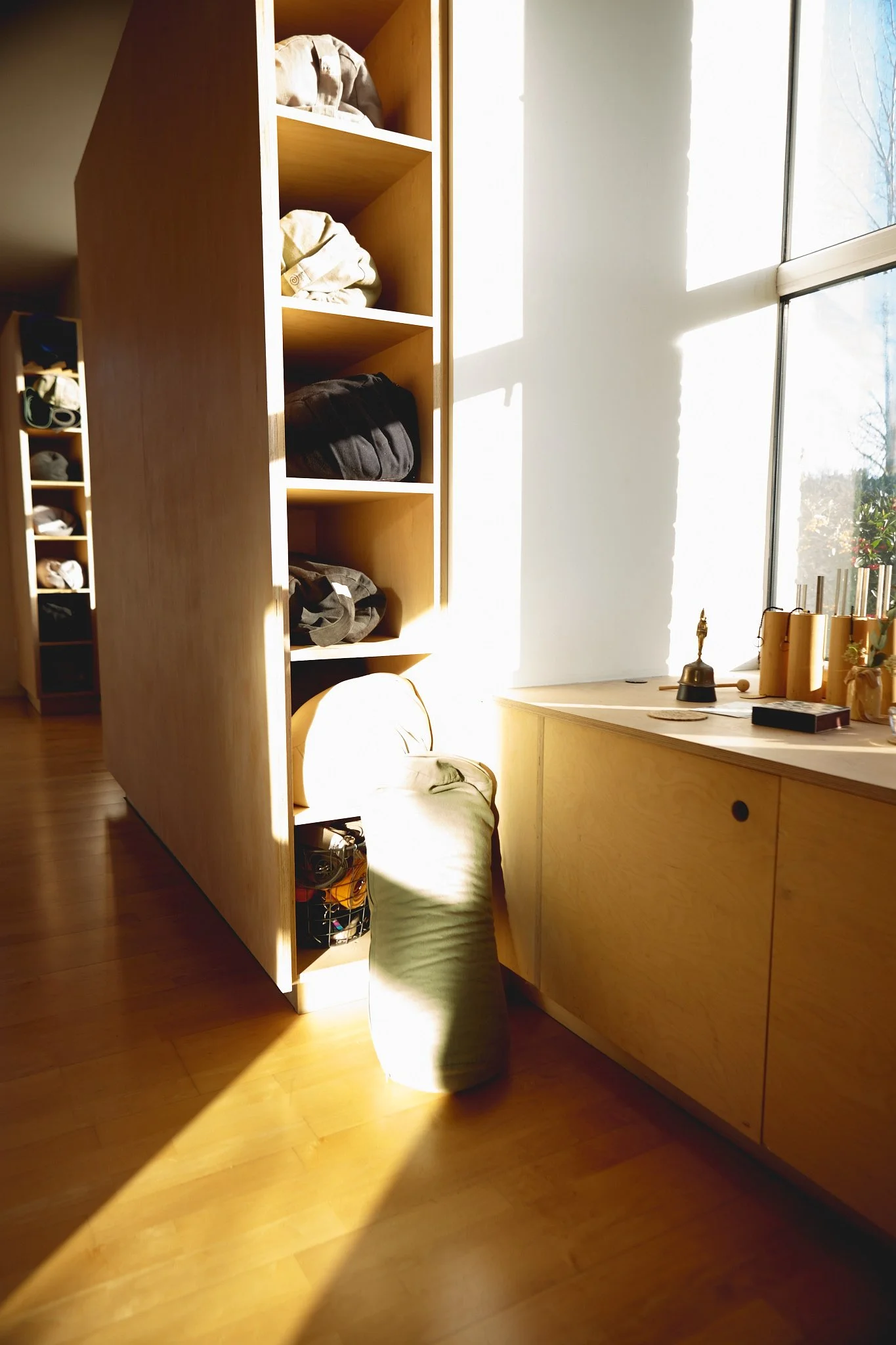 Sunlight streaming through a large window illuminating a wooden shelf filled with pilates equipment and a green yoga pillow resting on the floor nearby.