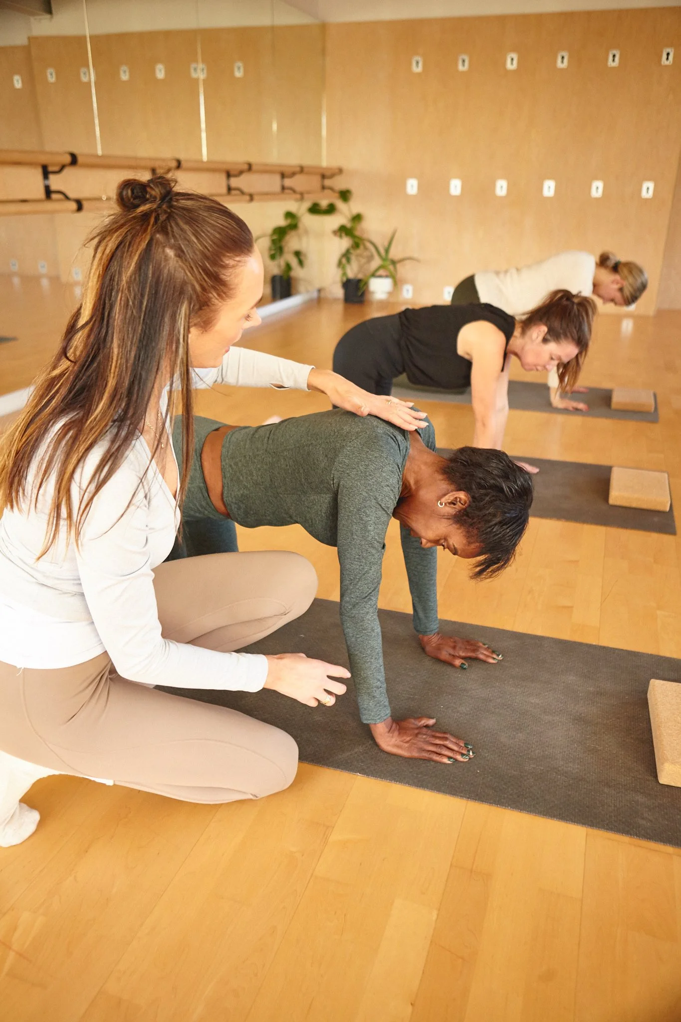 A yoga instructor assists a woman in a plank position during a Pilates class with three women on yoga mats in a studio.