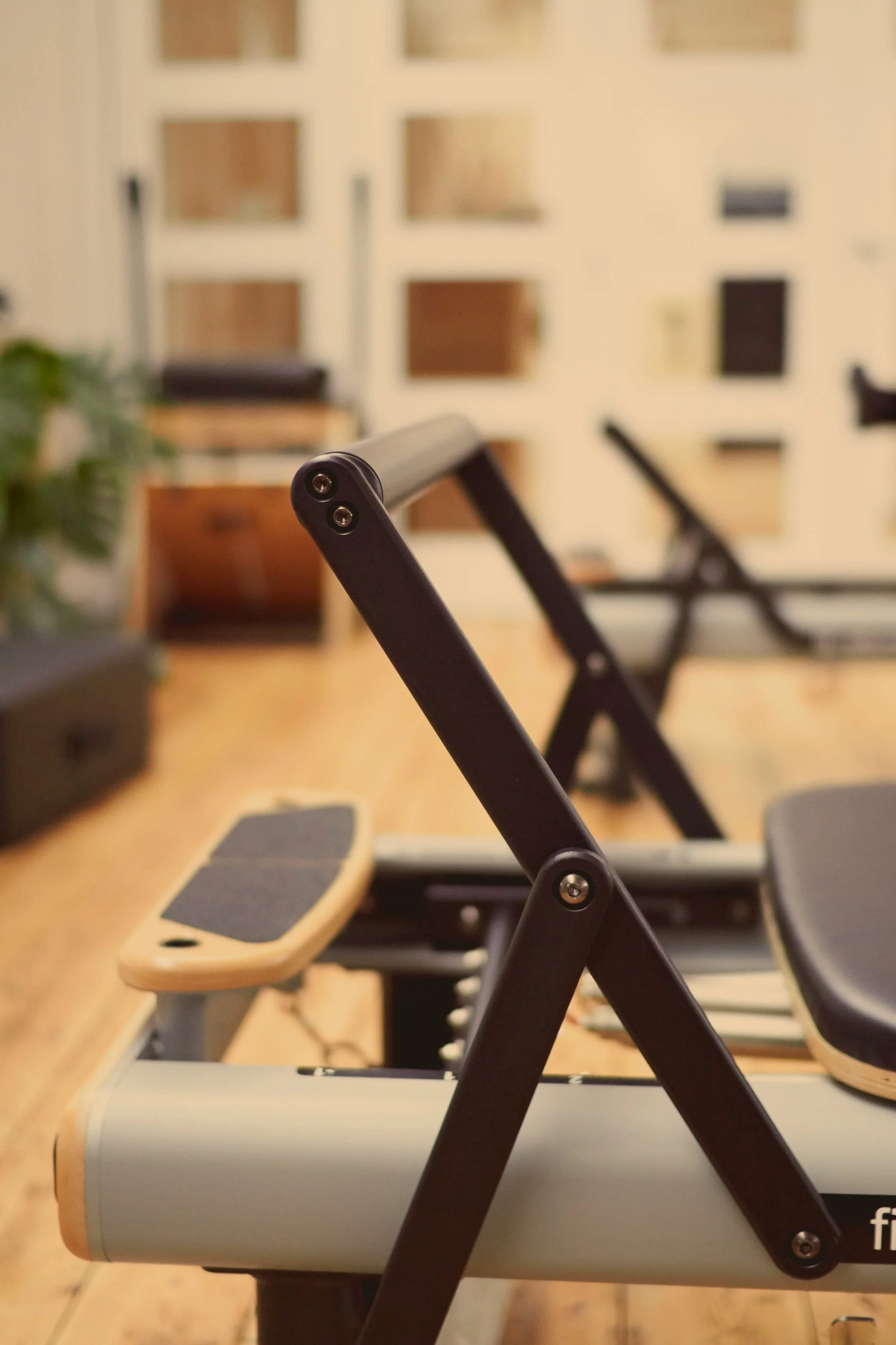 Close-up of the side of a Pilates reformer machine with a padded footrest in focus and a black padded seat in the background, in a room with wooden floors and beige walls.