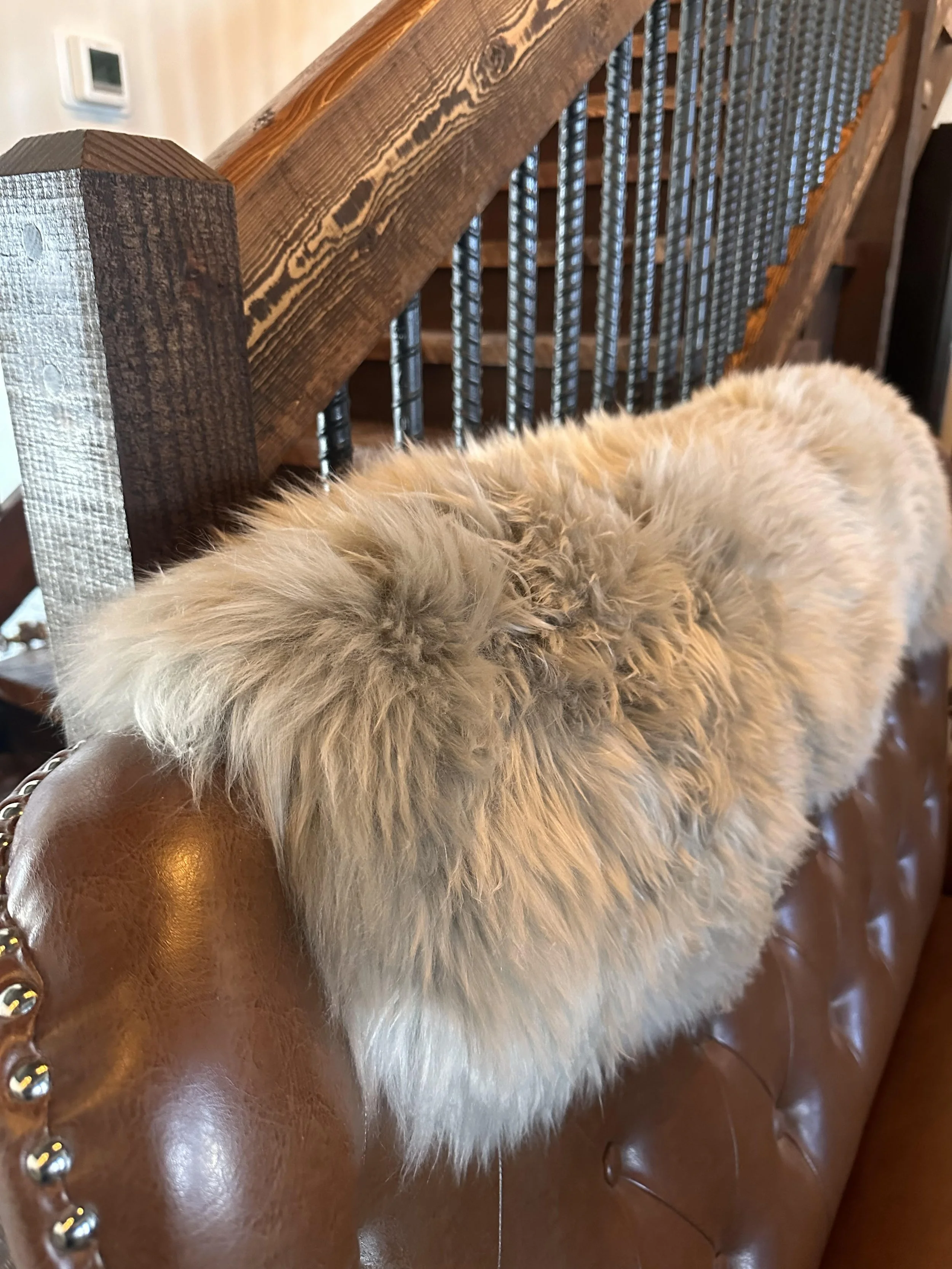 A fluffy, light-colored sheepskin resting on a brown leather couch near a wooden staircase.