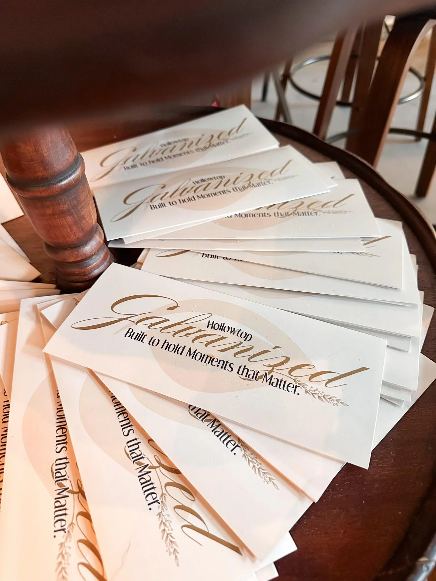 Multiple white paper cards with the words "Hollowtop Galvanized Built to hold Moments that Matter." printed in elegant gold and black font, spread out on a wooden surface near a wooden chair.