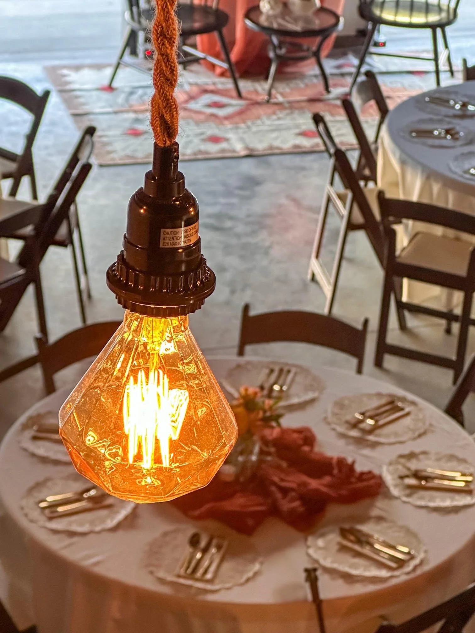 Close-up of a vintage-style Edison bulb chandelier hanging above a dining table with a red tablecloth and floral centerpiece, set for a meal in a rustic or industrial-style restaurant or event space.
