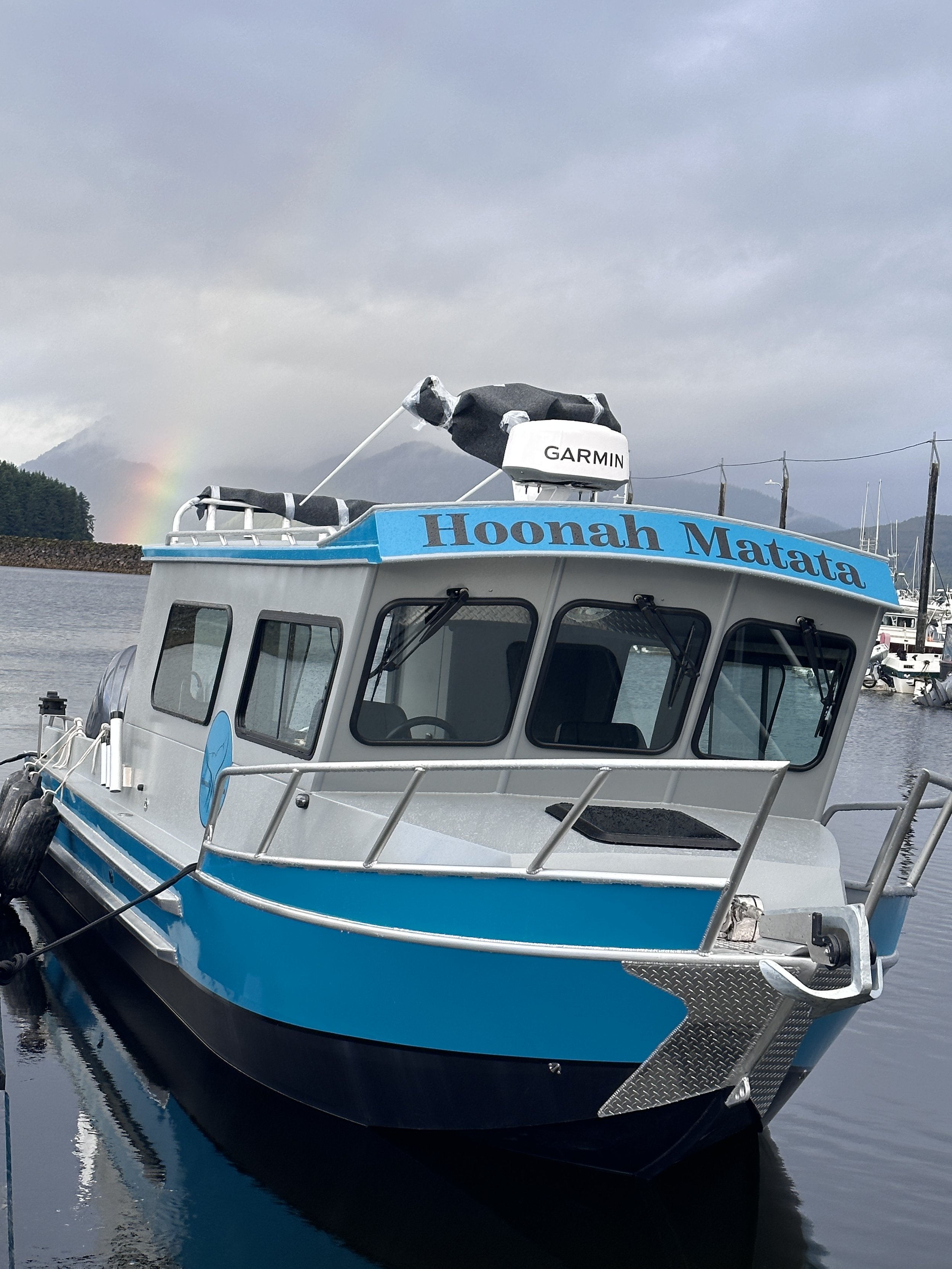 A boat named 'Hoonah Matata' docked at a marina, with mountains and a rainbow in the background.  Hoonah Matata will take guests whale watching and cruising quiet inlets, maybe pick up a crab pot too.