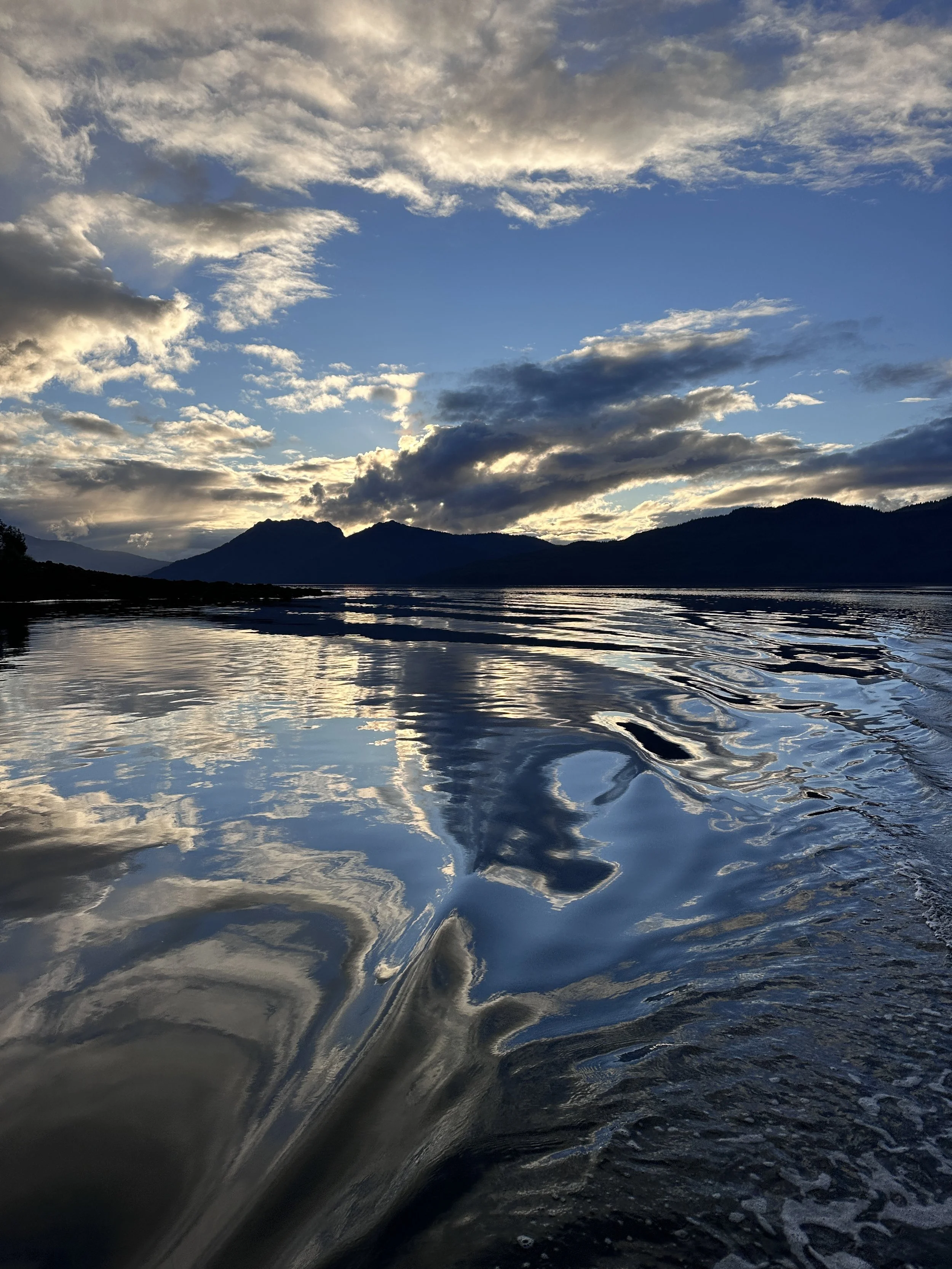 A scenic view of a lake with rippling water, surrounded by mountains under a partly cloudy sky during sunset or sunrise.