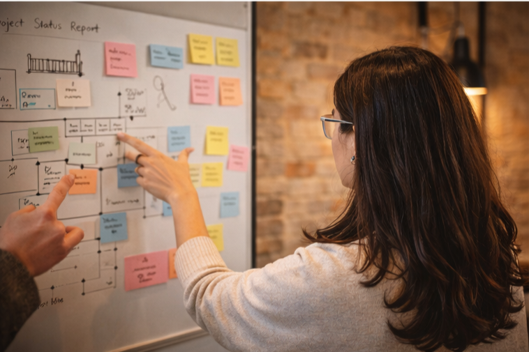 A woman and a man discussing a project status report chart on a whiteboard with colored sticky notes in a modern office with brick walls.