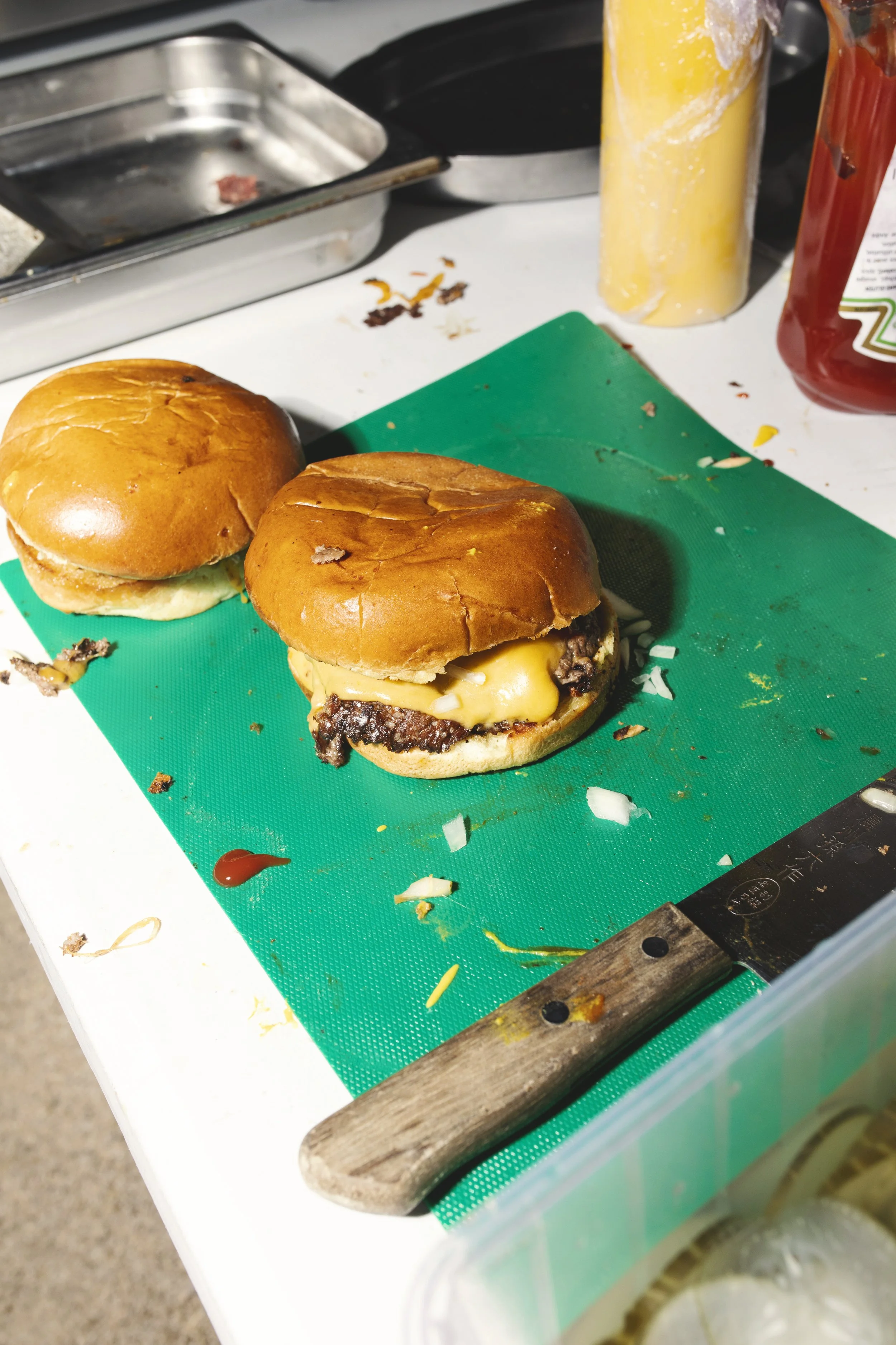 Deux cheeseburgers avec buns, fromage fondu, et viande, posés sur une planche en plastique verte sur une surface de travail. Un couteau à pain en bois est également visible.