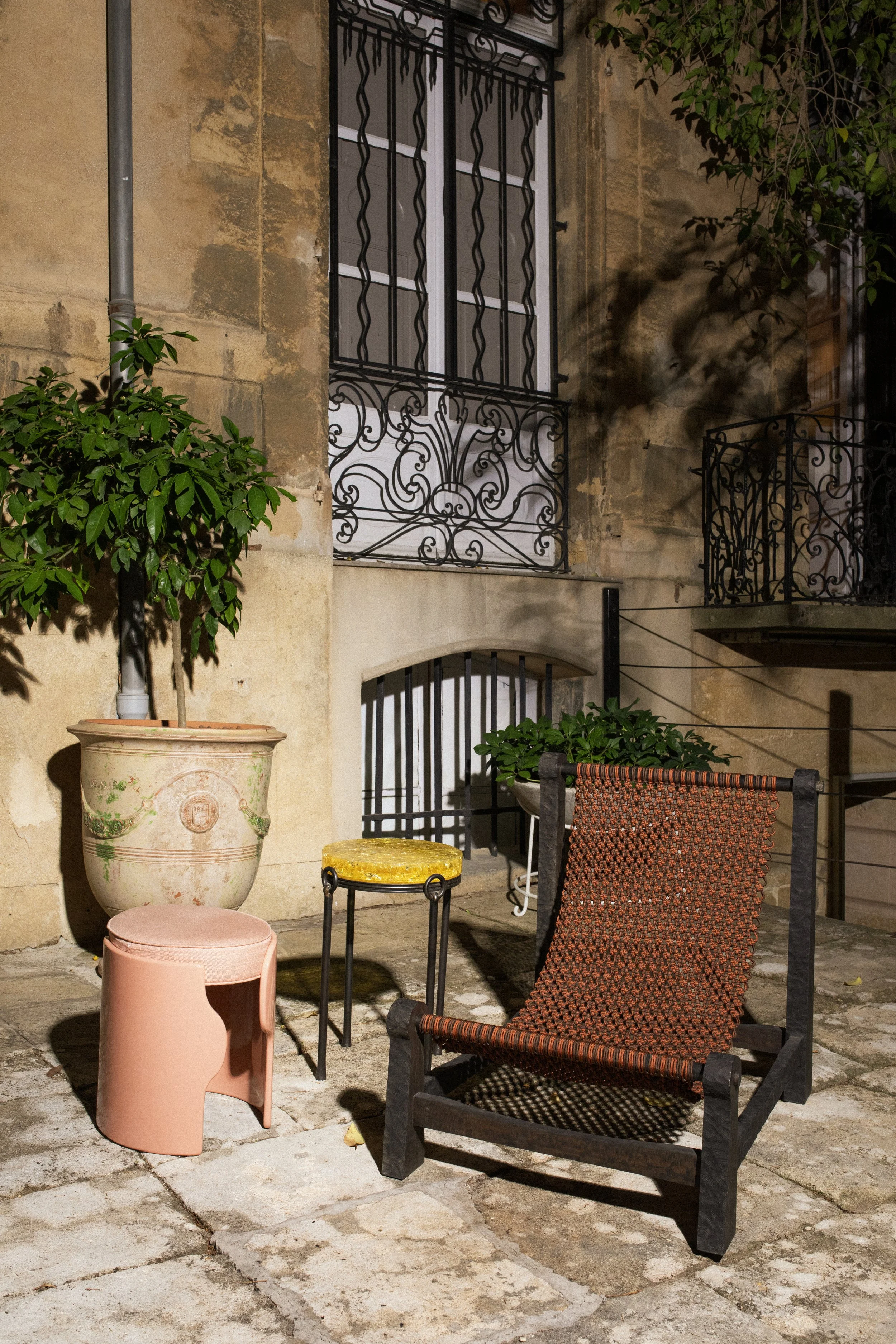 Une scène de cour extérieure la nuit, avec un fauteuil en bois et corde, une petite table jaune, un tabouret rose, une grande poterie avec un arbre, et un mur en pierre avec fenêtres à grille en fer forgé.
