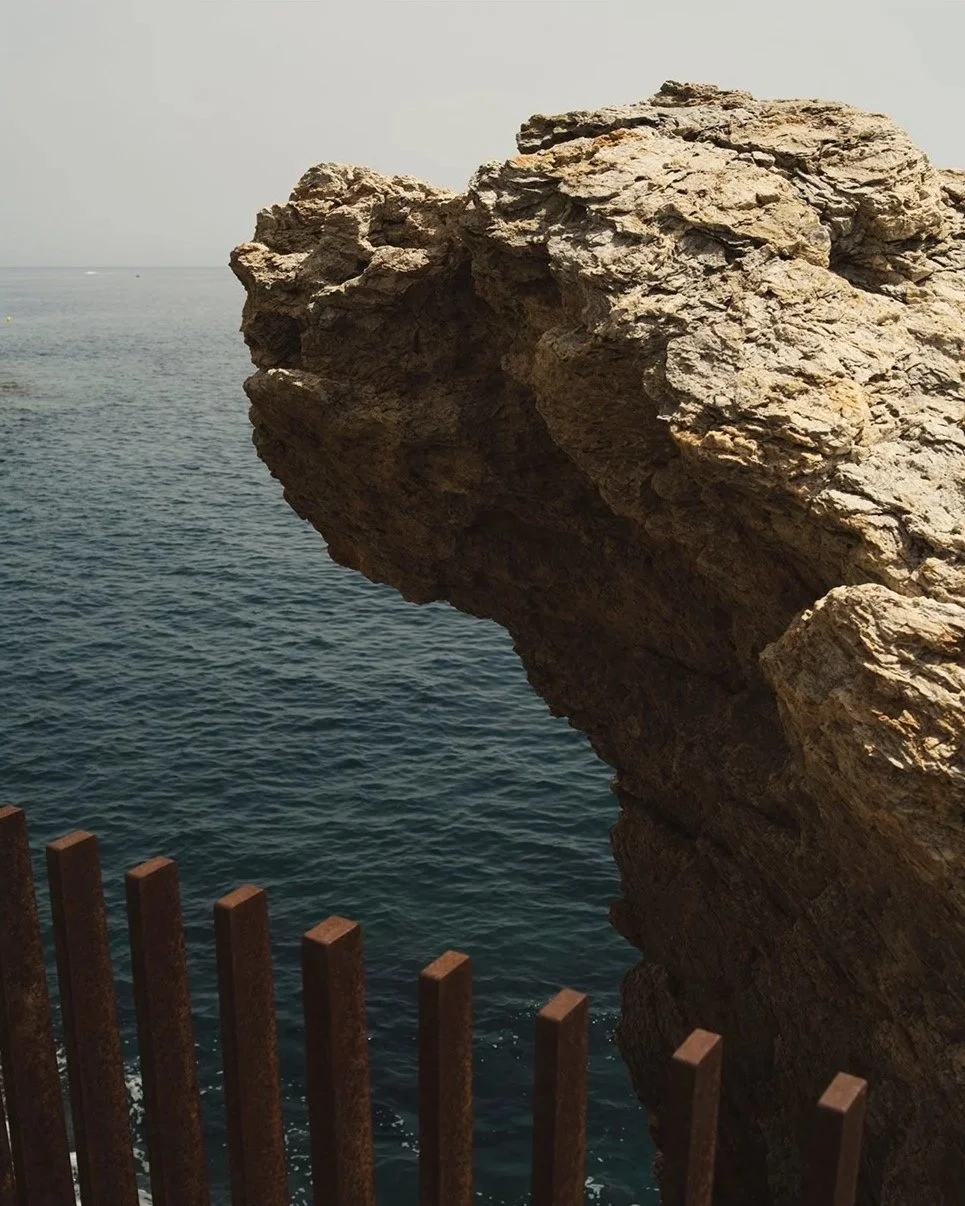Une grande roche s'avançant au-dessus de la mer, vue depuis une terrasse avec une barrière en fer rouillé.