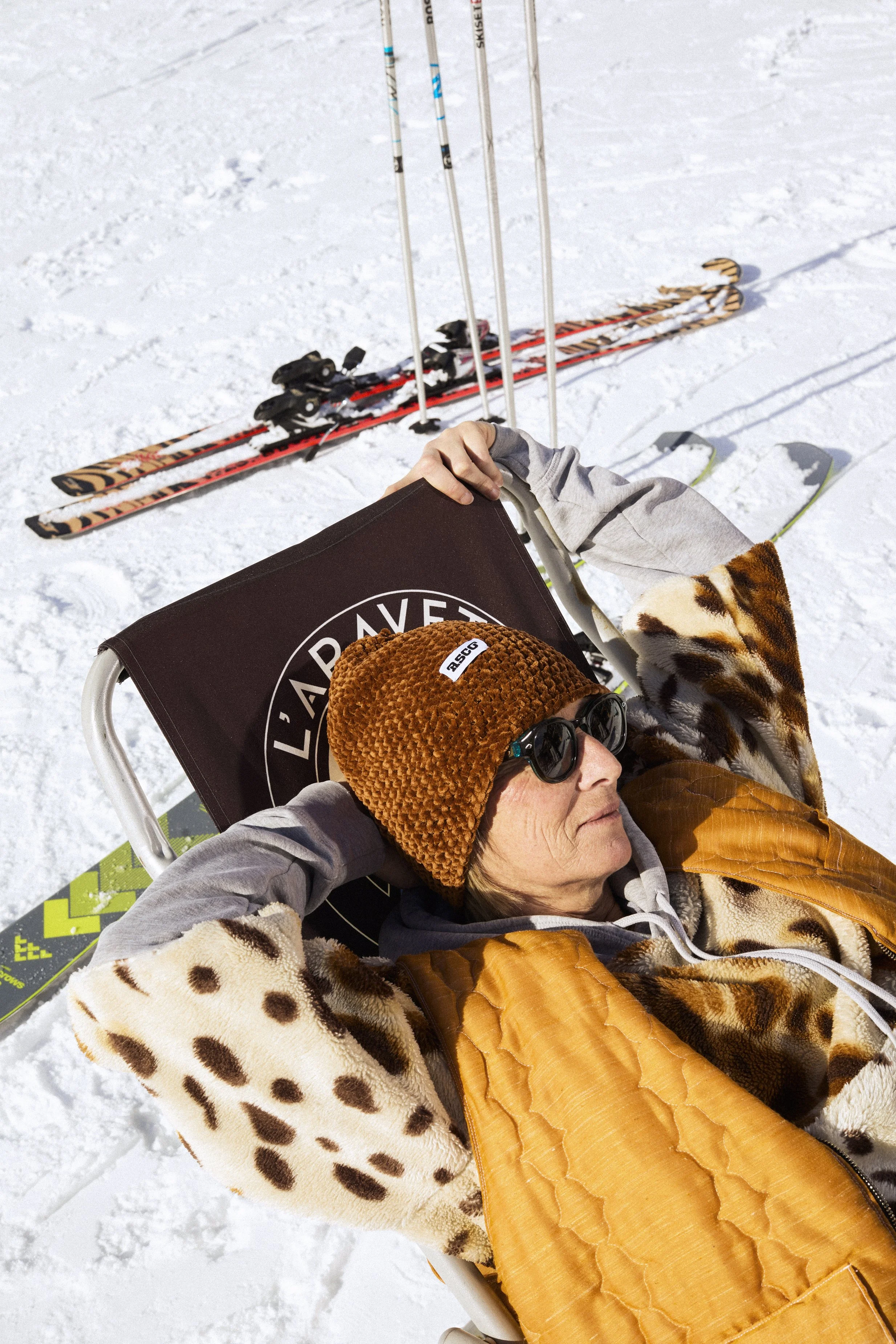 Une personne allongée sur une chaise longue sur la neige, portant des lunettes de soleil, un bonnet marron, une veste en fausse fourrure, des couvertures, avec des skis en arrière-plan.