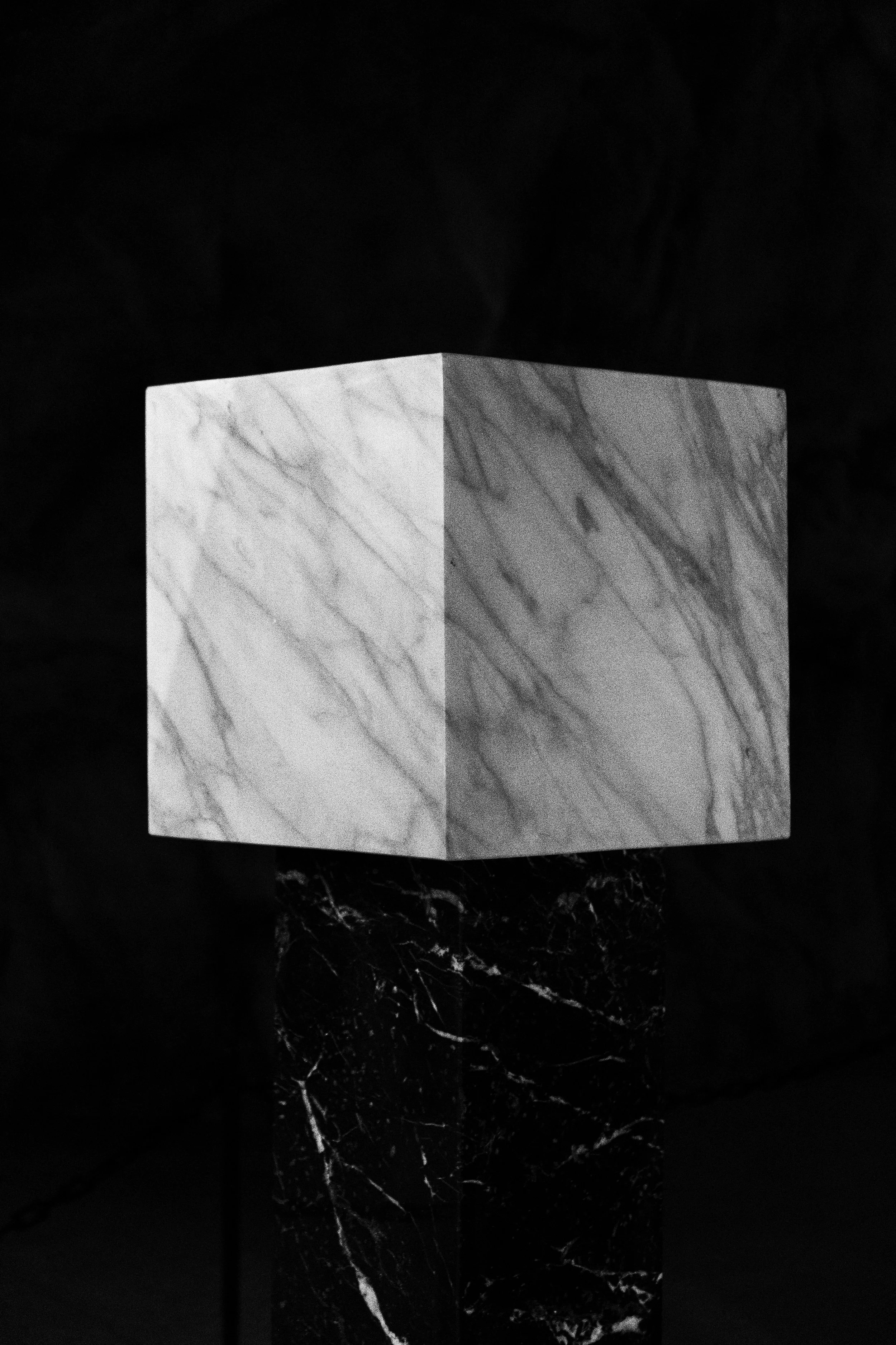 A white marble cube supported by a dark marble base, set against a black background.