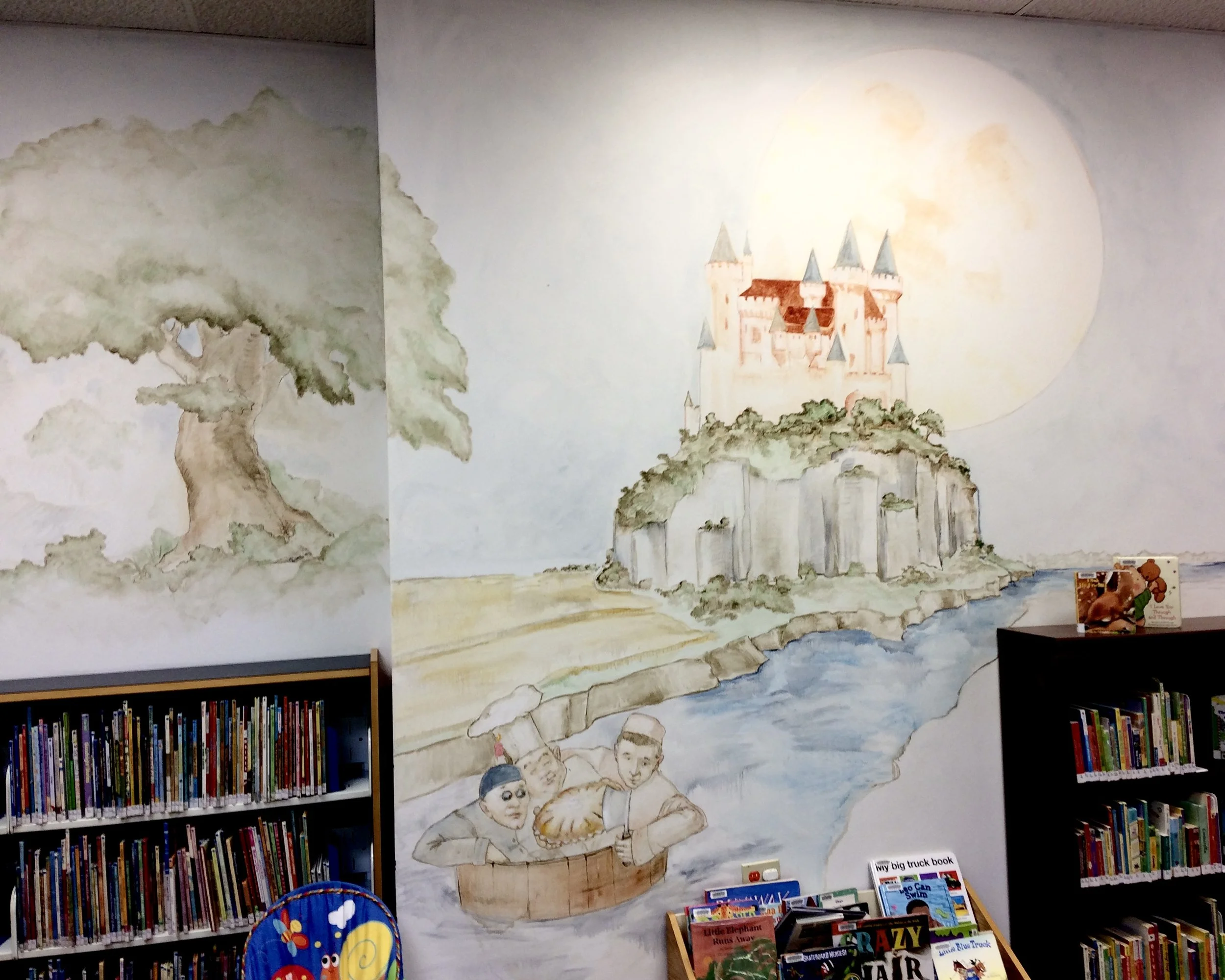 A mural on a wall depicting a whimsical castle with multiple towers, surrounded by trees and a hill. Below the castle, children are illustrated holding a large pie, with bookshelves and children’s books visible in the foreground.