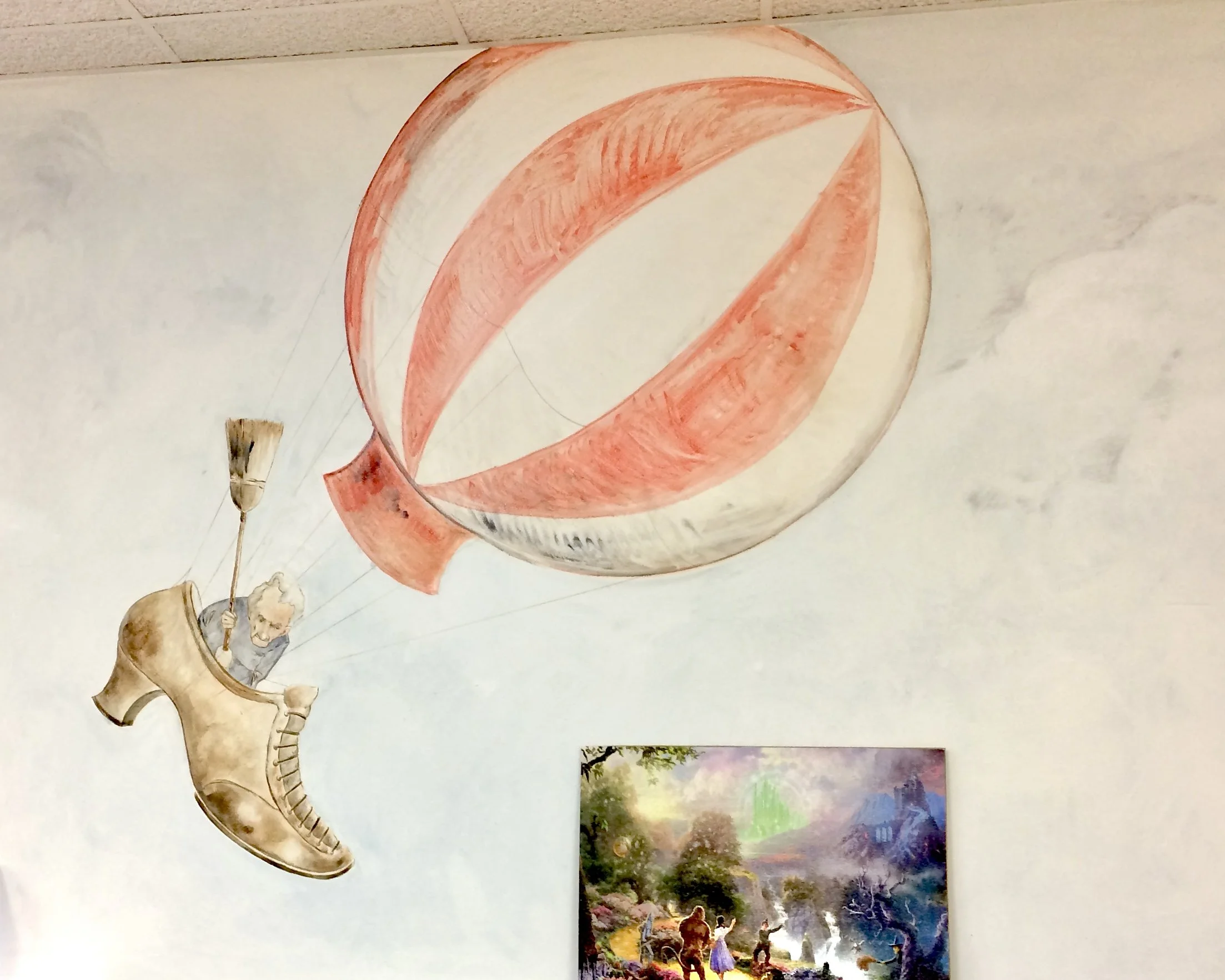 A child's drawing of a person riding a hot air balloon with the string attached to the basket, along with three landscape paintings on a white wall.
