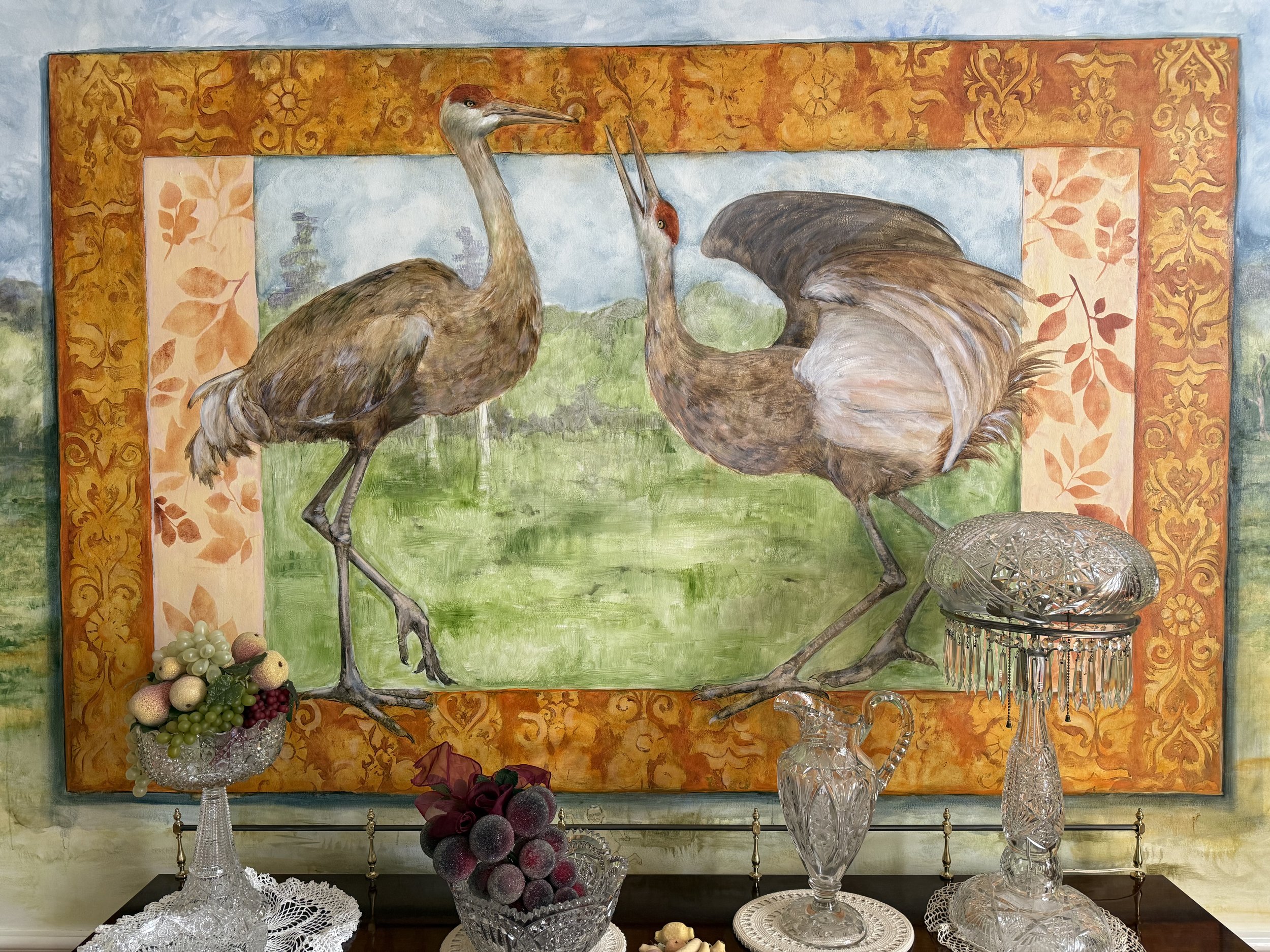Painting of two cranes inside an ornate gold and pink frame, with various glass and crystal decorative items and fruit in front.