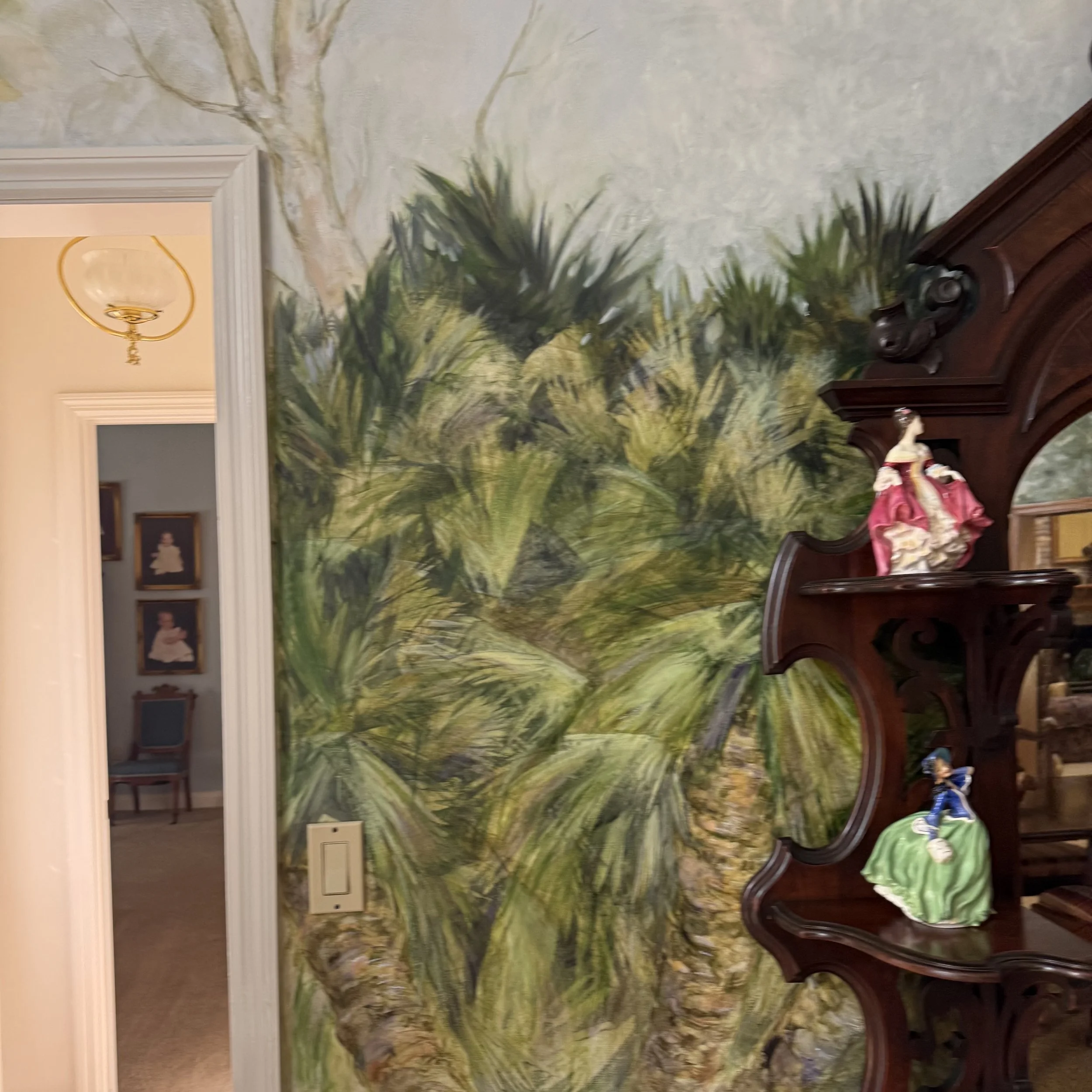 A mural of a green, wooded landscape with trees and grass painted on a wall in a room. There is a doorway and framed pictures visible to the left, and a wooden piece of furniture with decorative items on it on the right side.