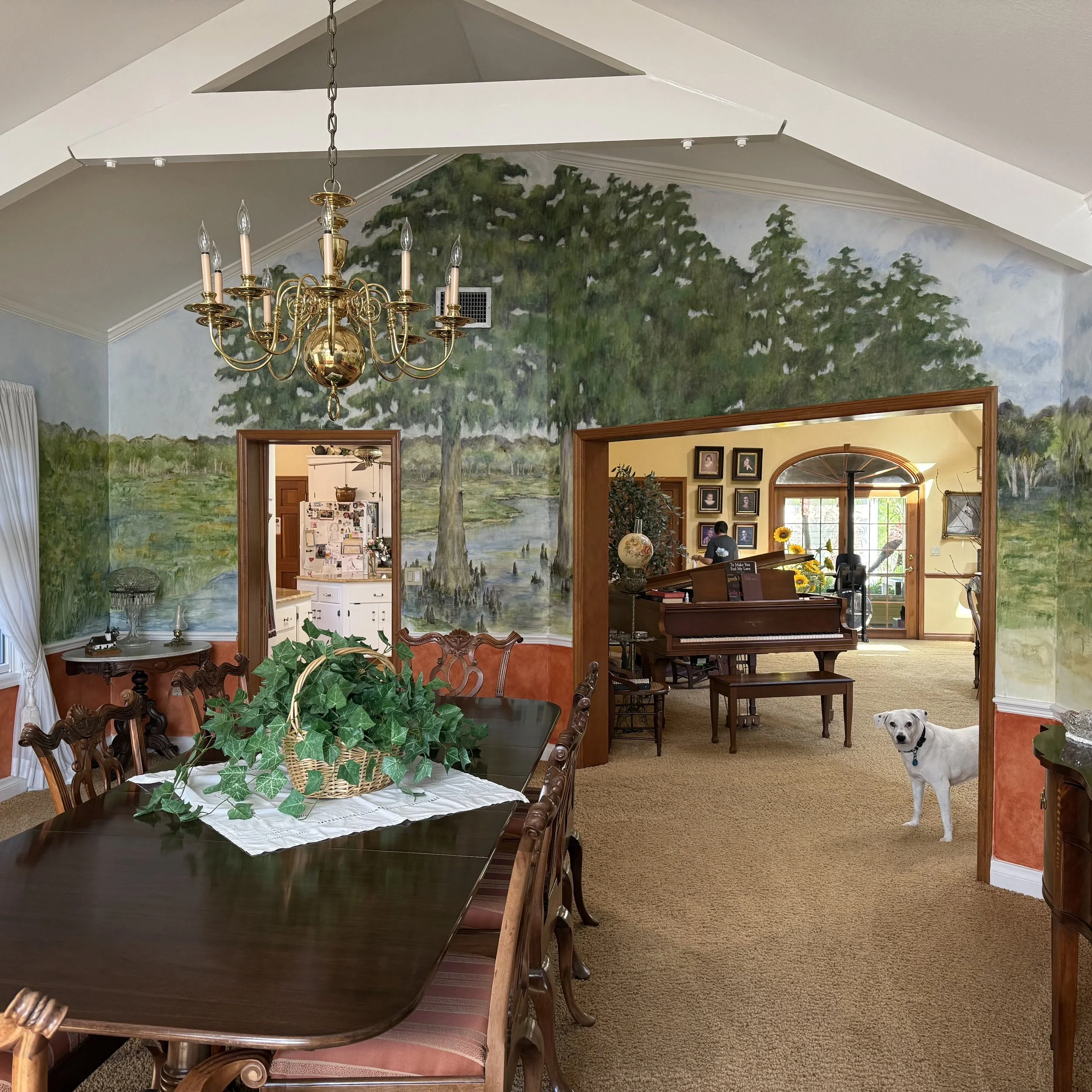 Dining room with a large wooden table, ornate chairs, a chandelier hanging from the ceiling, and a wall mural of trees and a pond. There is a small dog standing on the carpeted floor near the doorway.