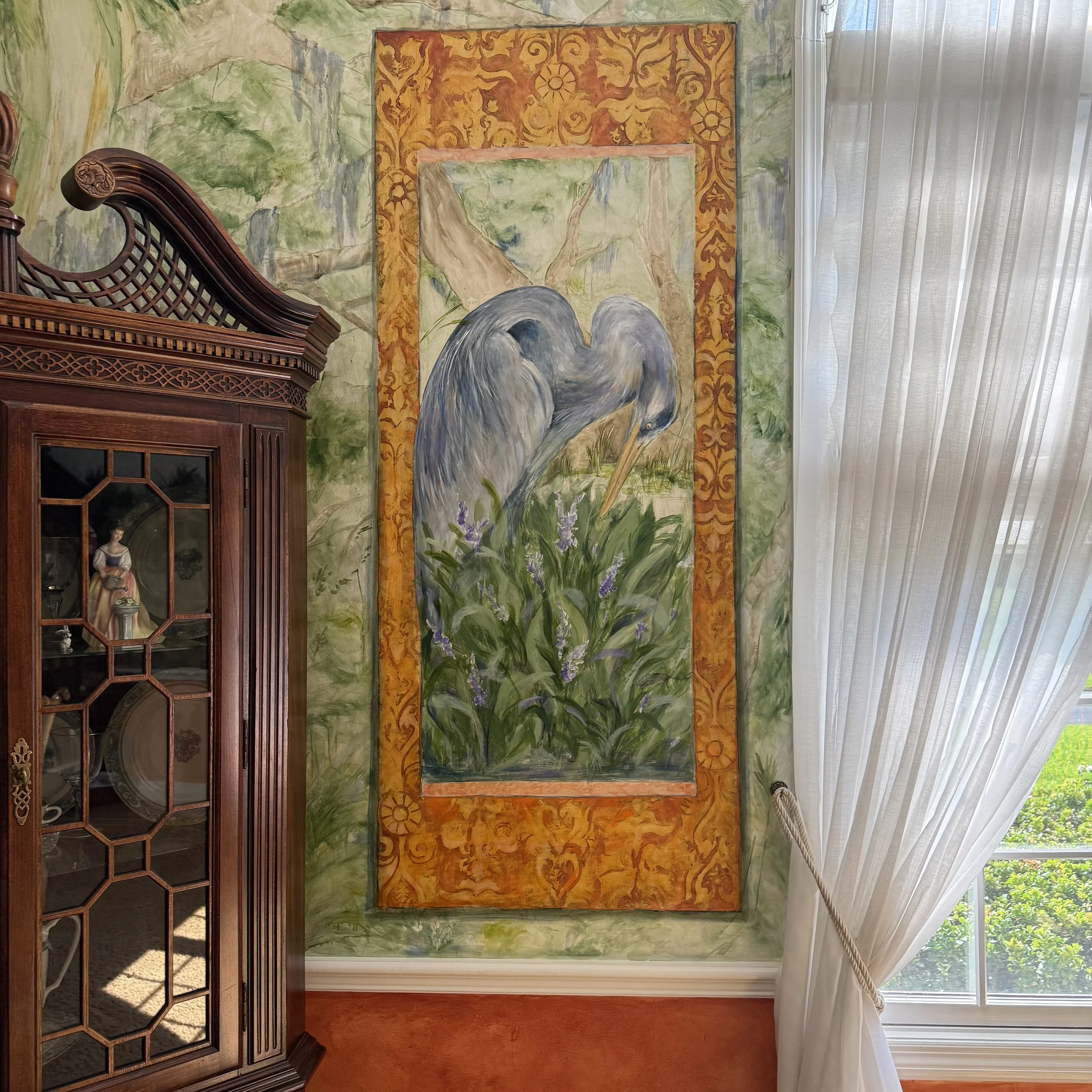 Wall mural depicting a scene of a heron standing among tall grass and purple flowers, framed by a decorative red and gold border, beside a window with sheer white curtains.