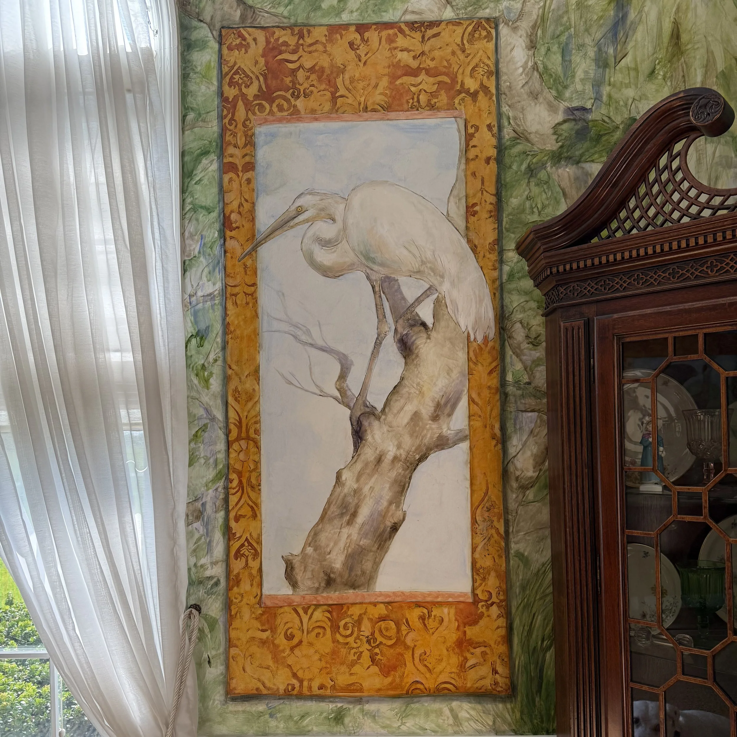 A large framed painting of a white heron bird set against an orange and gold decorative background, hanging on a wall with a green, nature-themed mural. The scene is illuminated by natural light coming from sheer white curtains on the left.