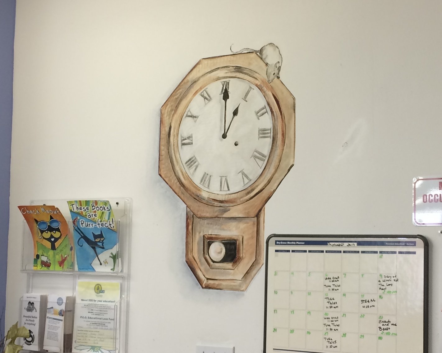 A wall clock with a wooden frame shows the time as 12:02. To the left, there are colorful children's books with cartoon cats on the covers and a brochure holder. To the right, a whiteboard monthly planner displays events and notes.