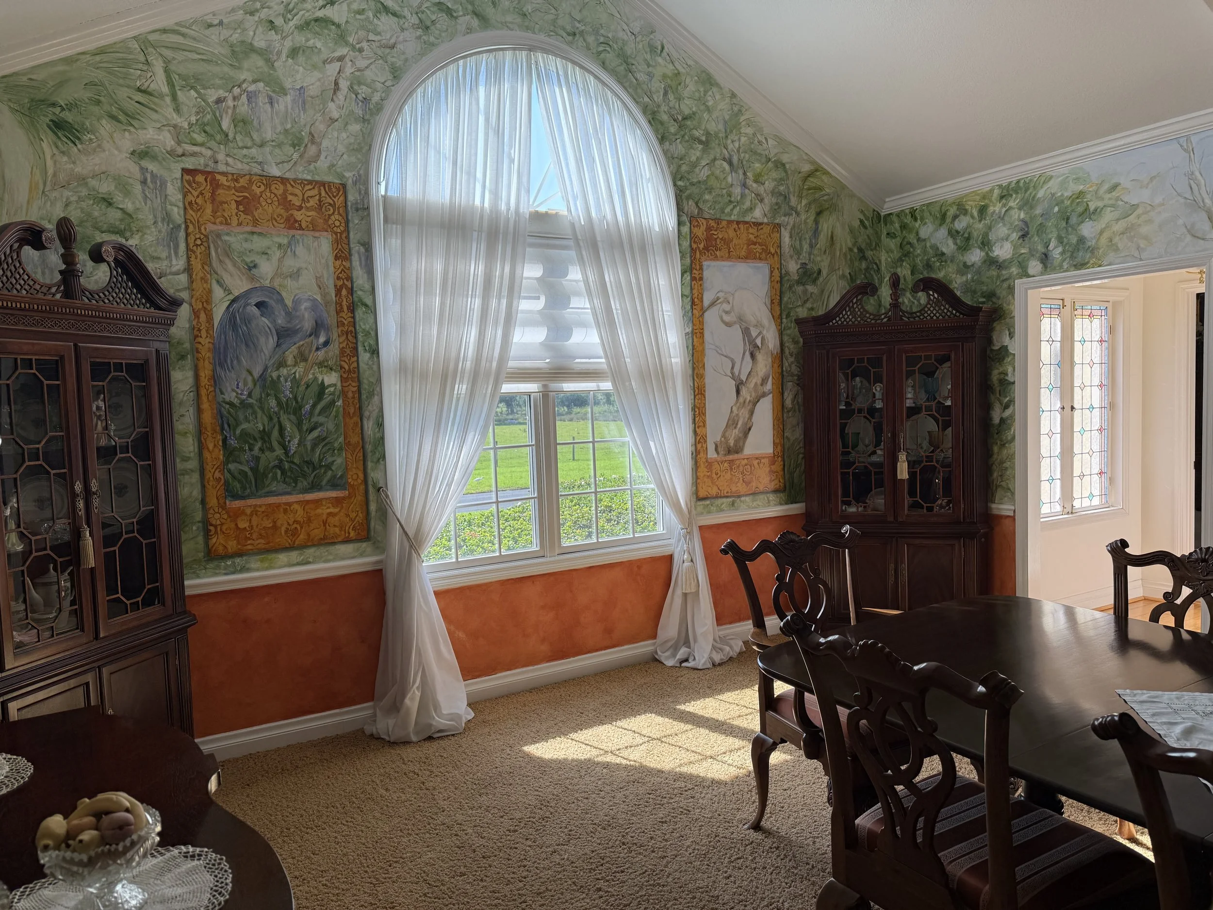 Interior of a dining room with large arched windows, white curtains, and wall murals of herons and trees. There are dark wooden china cabinets, a wooden dining table with matching chairs, a small table with a lace doily and decorative eggs, and a carpeted floor.
