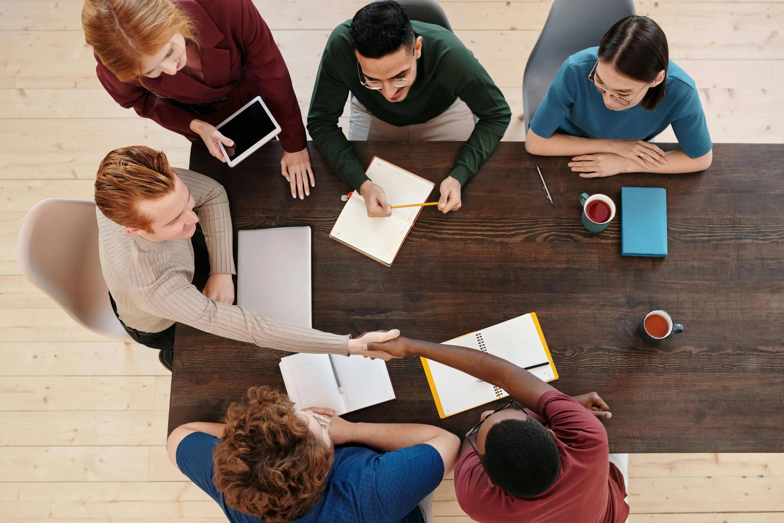 A diverse group of six people gathered around a wooden table in a meeting, shaking hands, with notebooks, laptops, and coffee cups on the table.