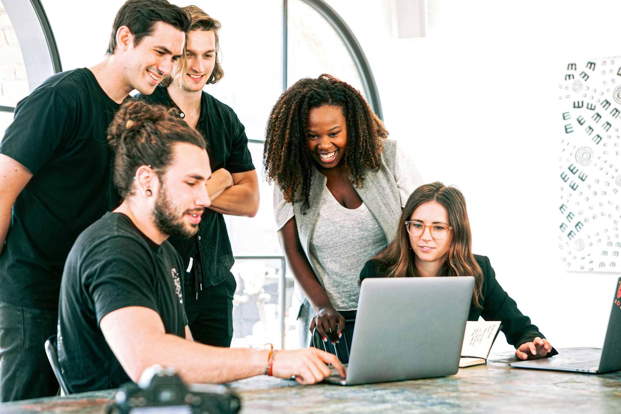 Five young adults, three men and two women, collaborate around a laptop in a modern, brightly lit office