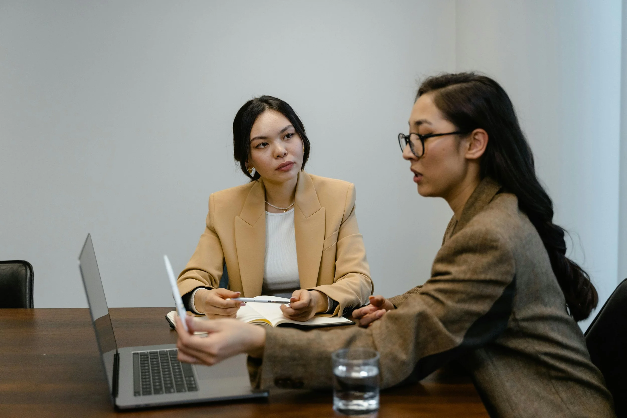 Two women in business attire having a meeting in a conference room, with a laptop and a glass of water on the table.