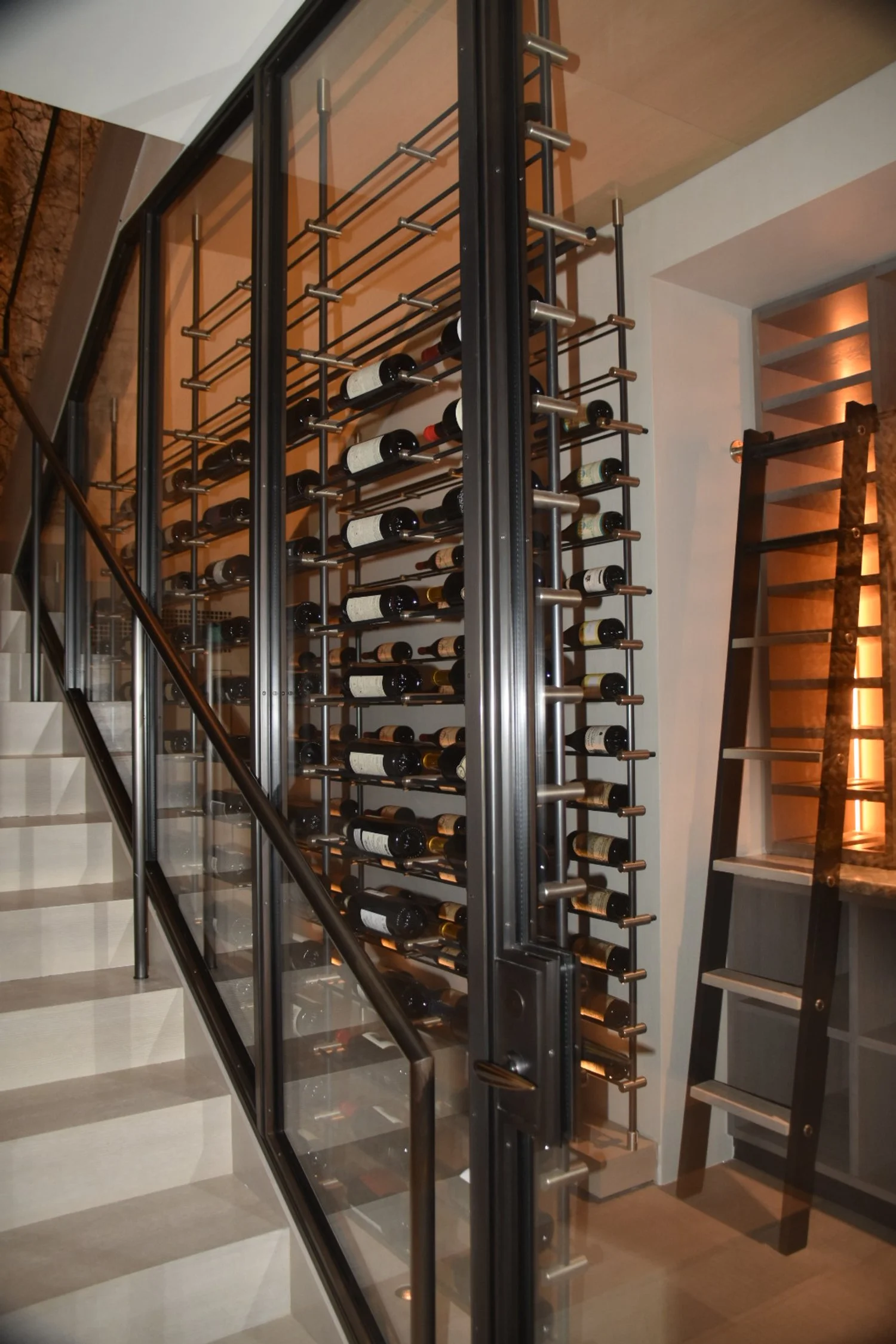 A wine cellar rack filled with bottles behind a glass door, next to a black ladder and staircase.